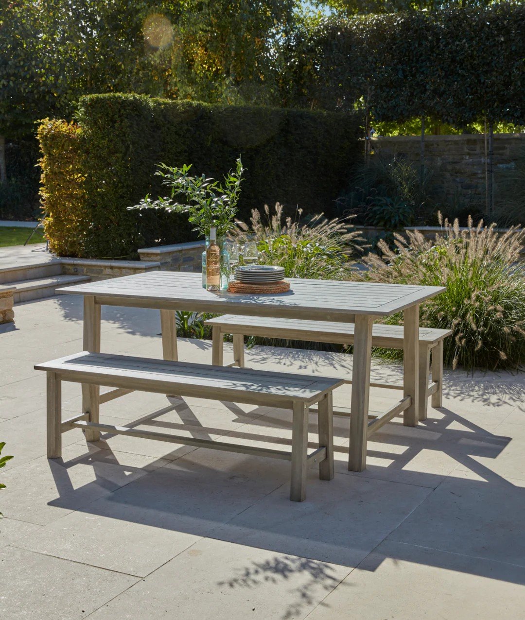 Outdoor patio with a white wooden dining table, a bench, and chairs, set with plates, glasses, and bottles, surrounded by lush greenery and plants.