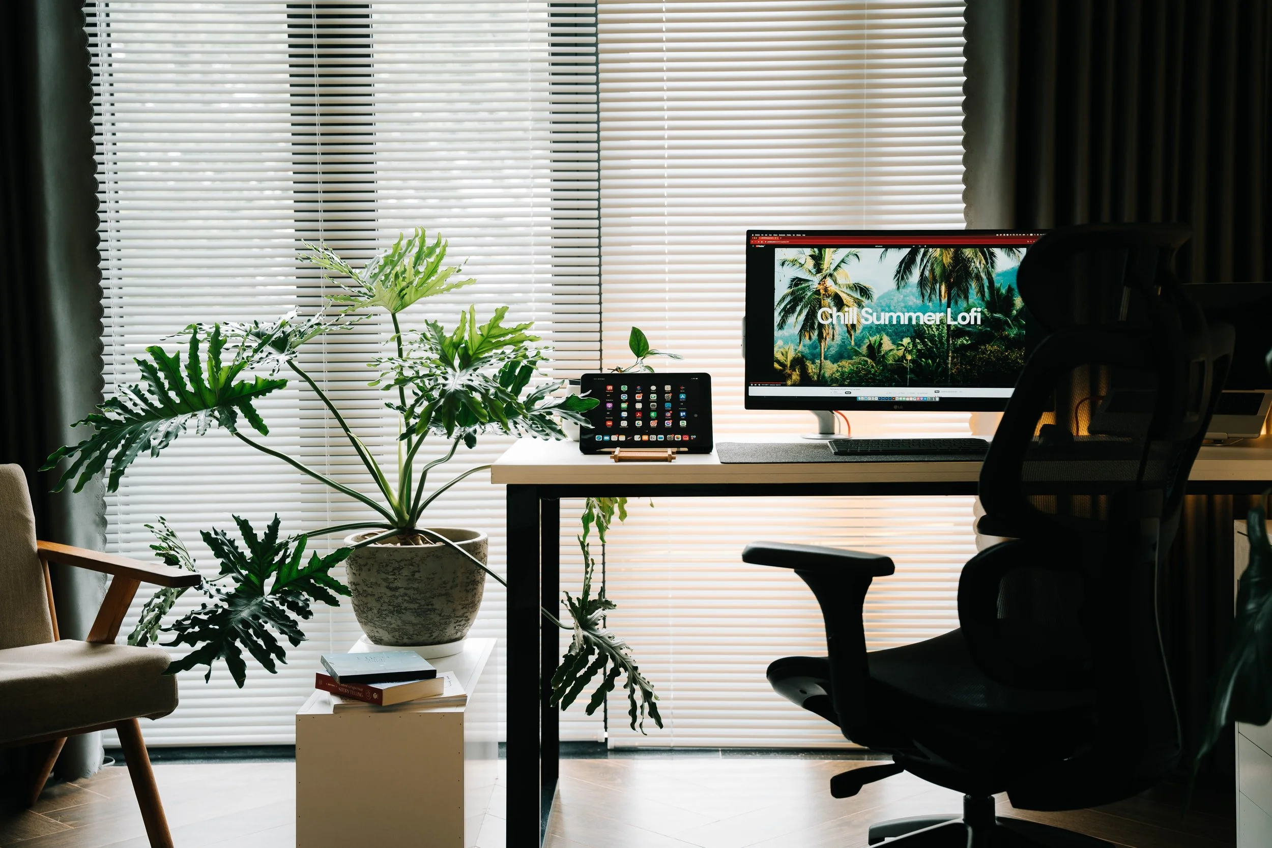 A modern home office with a large window covered by horizontal blinds, greenery on the desk, including a large potted plant and smaller plants, a monitor displaying a tropical scene with palm trees and the text 'Chill Summer Lofi', a tablet, books, and a chair.