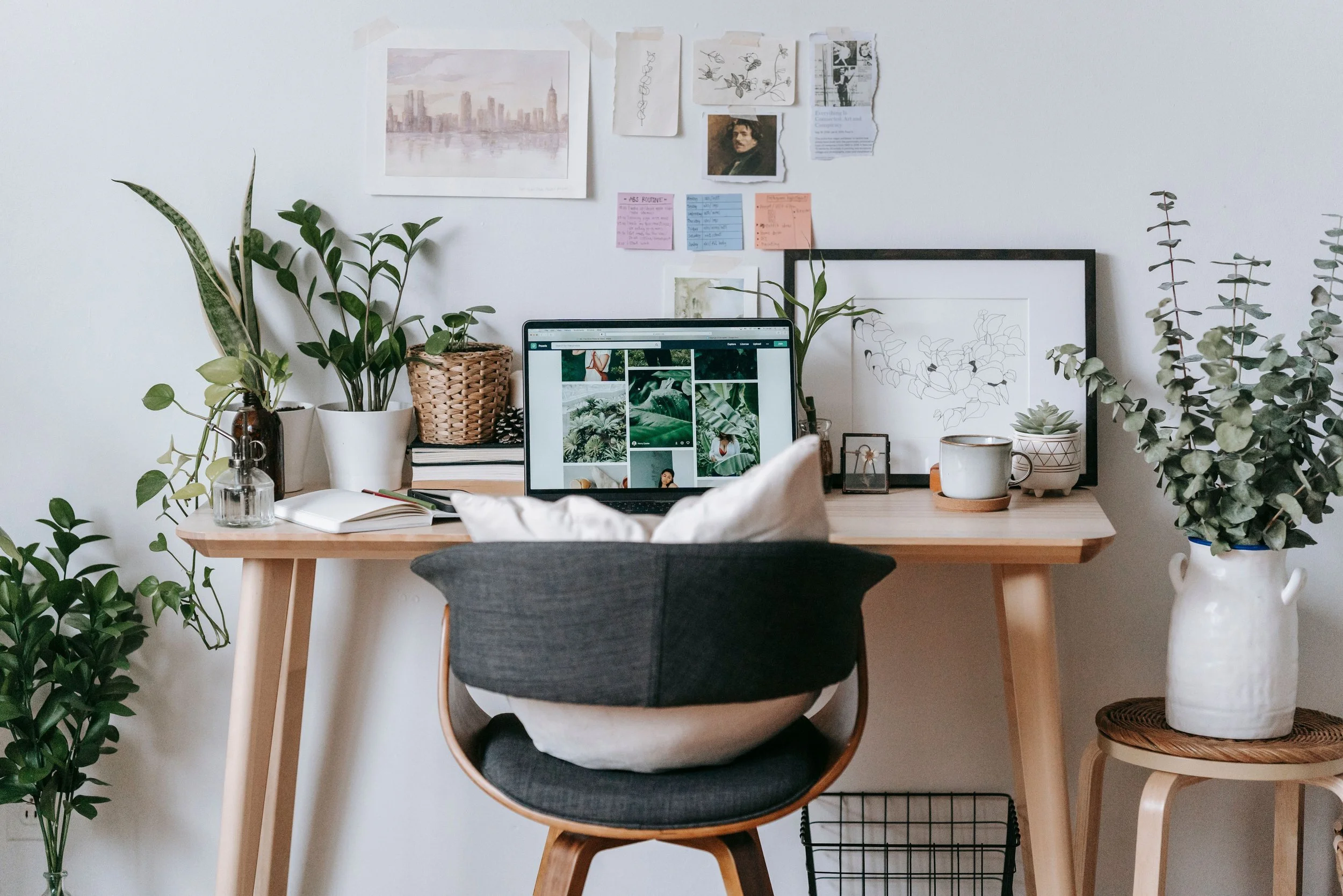 A home office with a wooden desk and an ergonomic chair with cushions. The desk has plants, a laptop displaying images of greenery, a notebook, and writing tools. The wall behind features framed artwork, sketches, photographs, and colorful sticky notes. Potted plants are arranged on the desk and nearby on a small table and the floor.