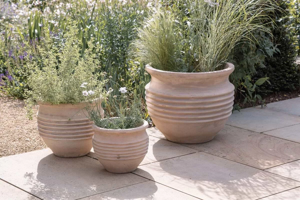 Three large off-white ceramic flower pots with horizontal ridges, containing green plants, on a tiled patio with shadows and garden plants in the background.