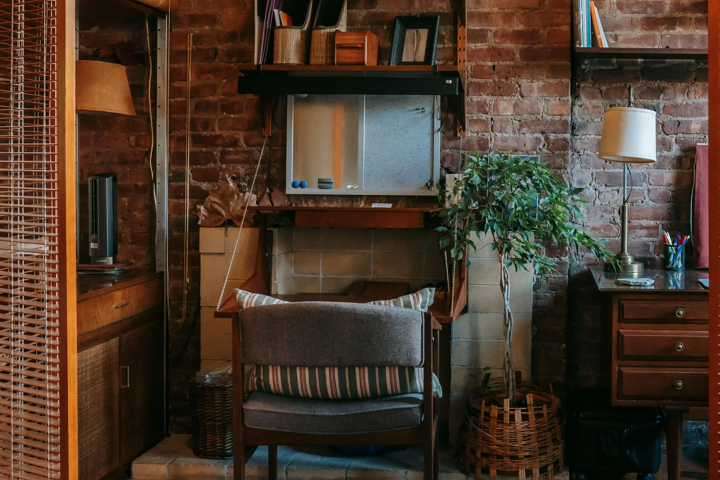 A cozy interior with an exposed brick wall, a mirror, and various decorative items, including woven baskets, a potted plant, and a standing lamp. A workspace with a small table and an upholstered chair with striped cushions is also visible.