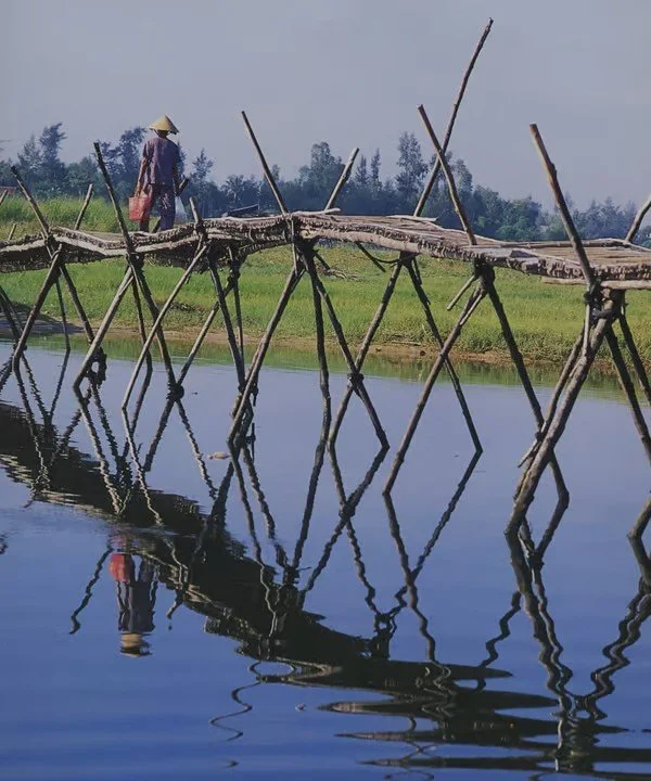 A person wearing a traditional conical hat and colorful clothing walking on a rustic bamboo bridge over a body of water in a rural area with greenery in the background.