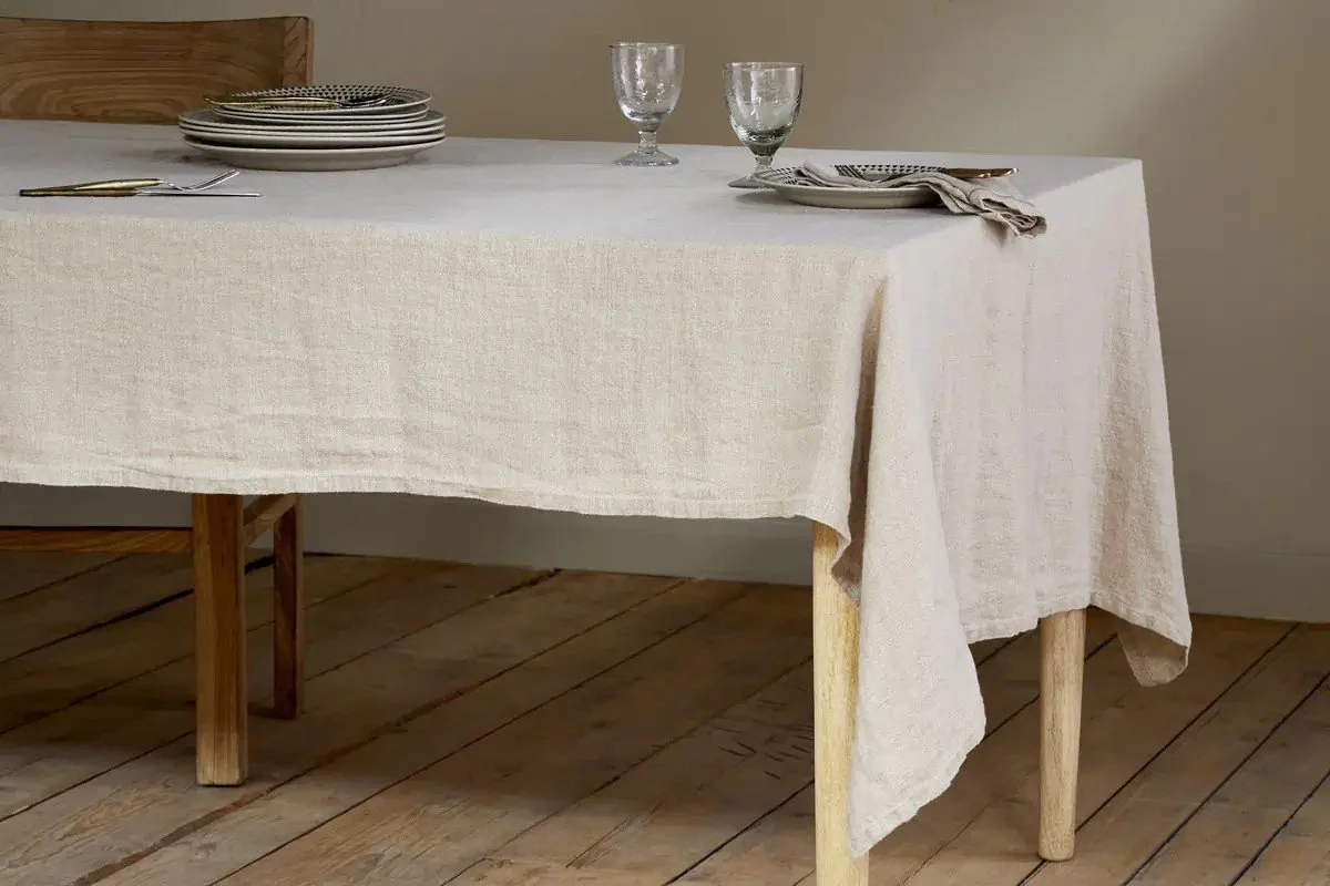 A dining table set with plates, glasses, and napkins, covered with a white tablecloth, in a room with wooden flooring and beige walls.