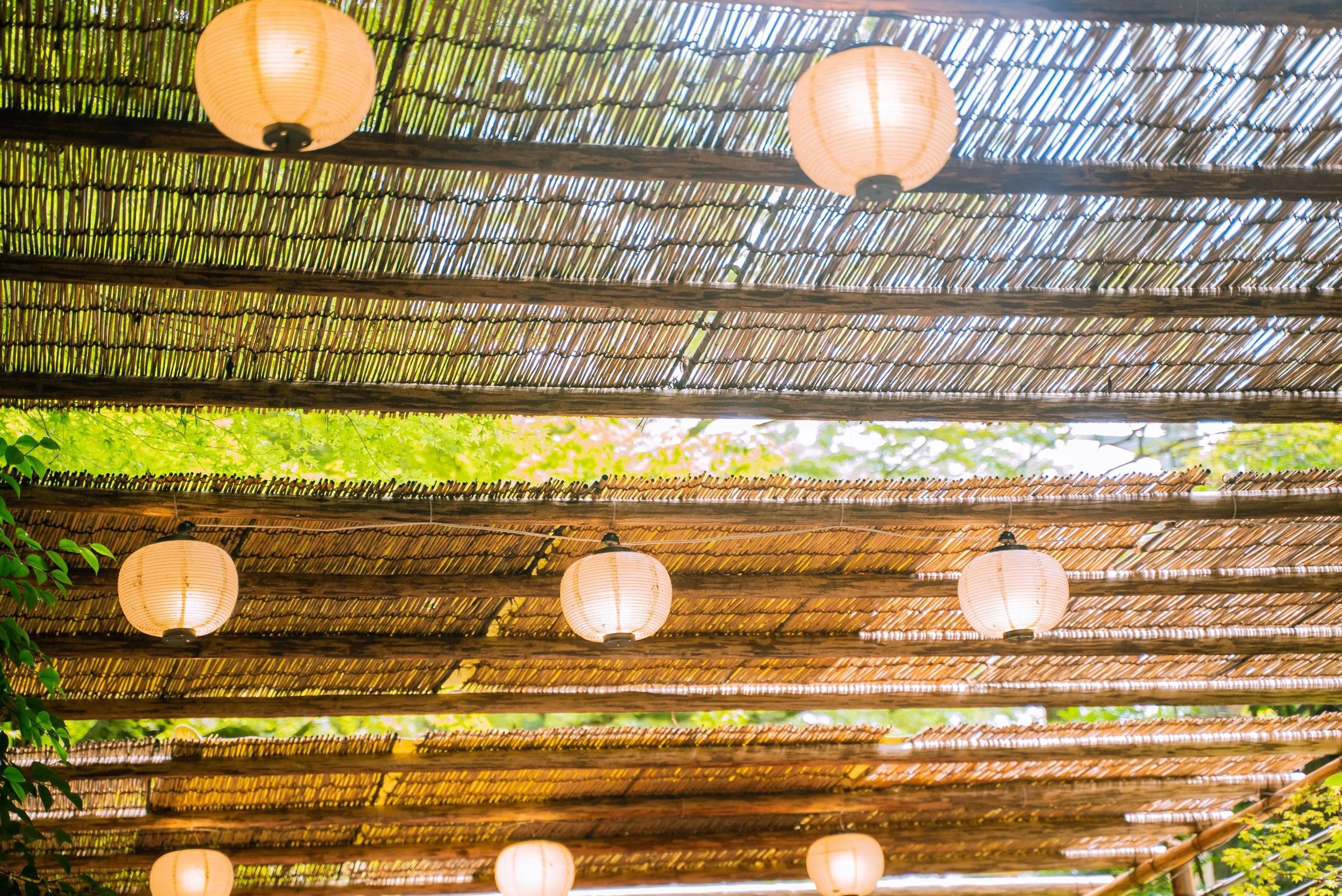 Japanese paper lanterns hanging from a bamboo and wooden pergola, with green leaves and sunlight filtering through the roof.