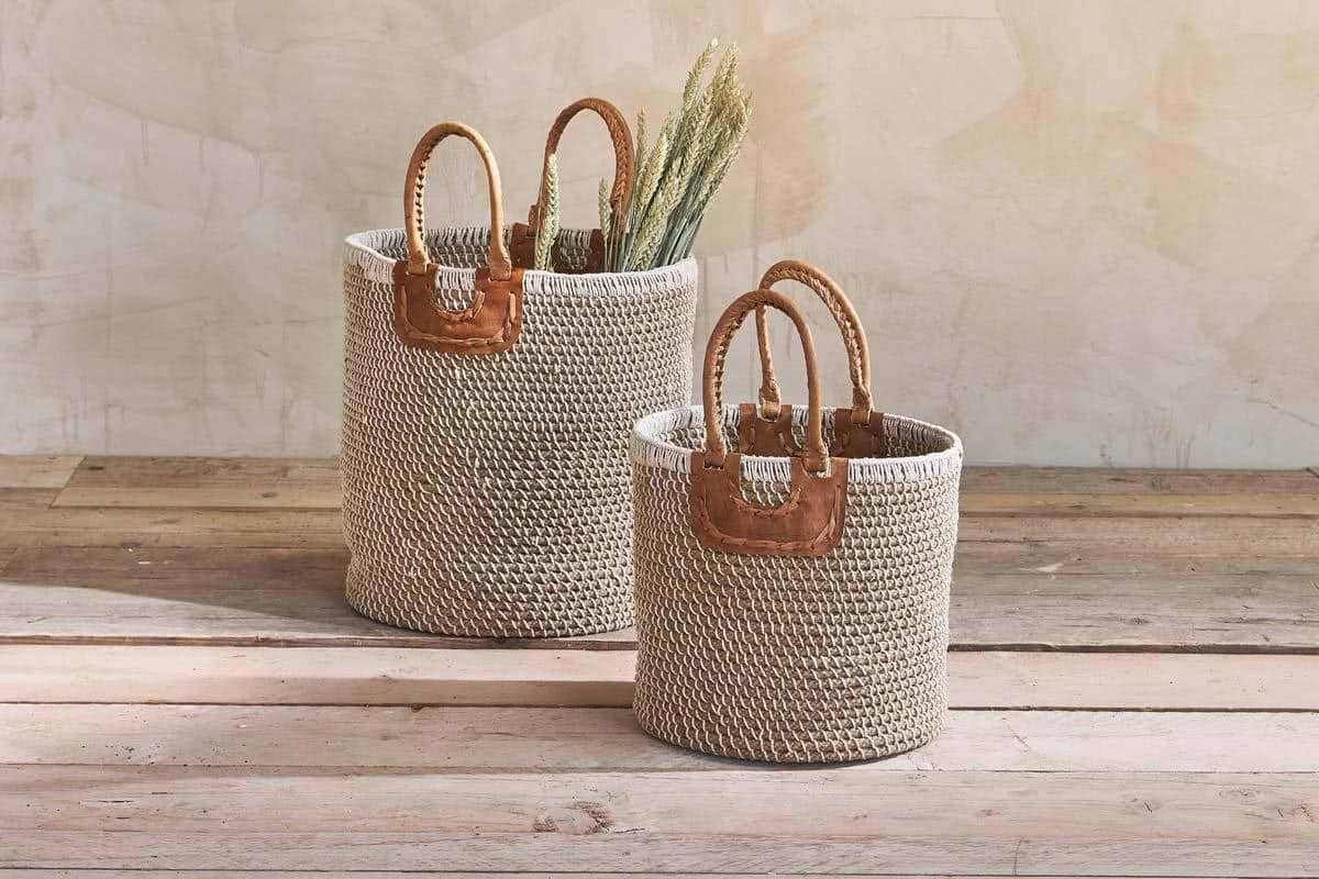 Two woven baskets with leather handles on a wooden surface, one containing dried plants.