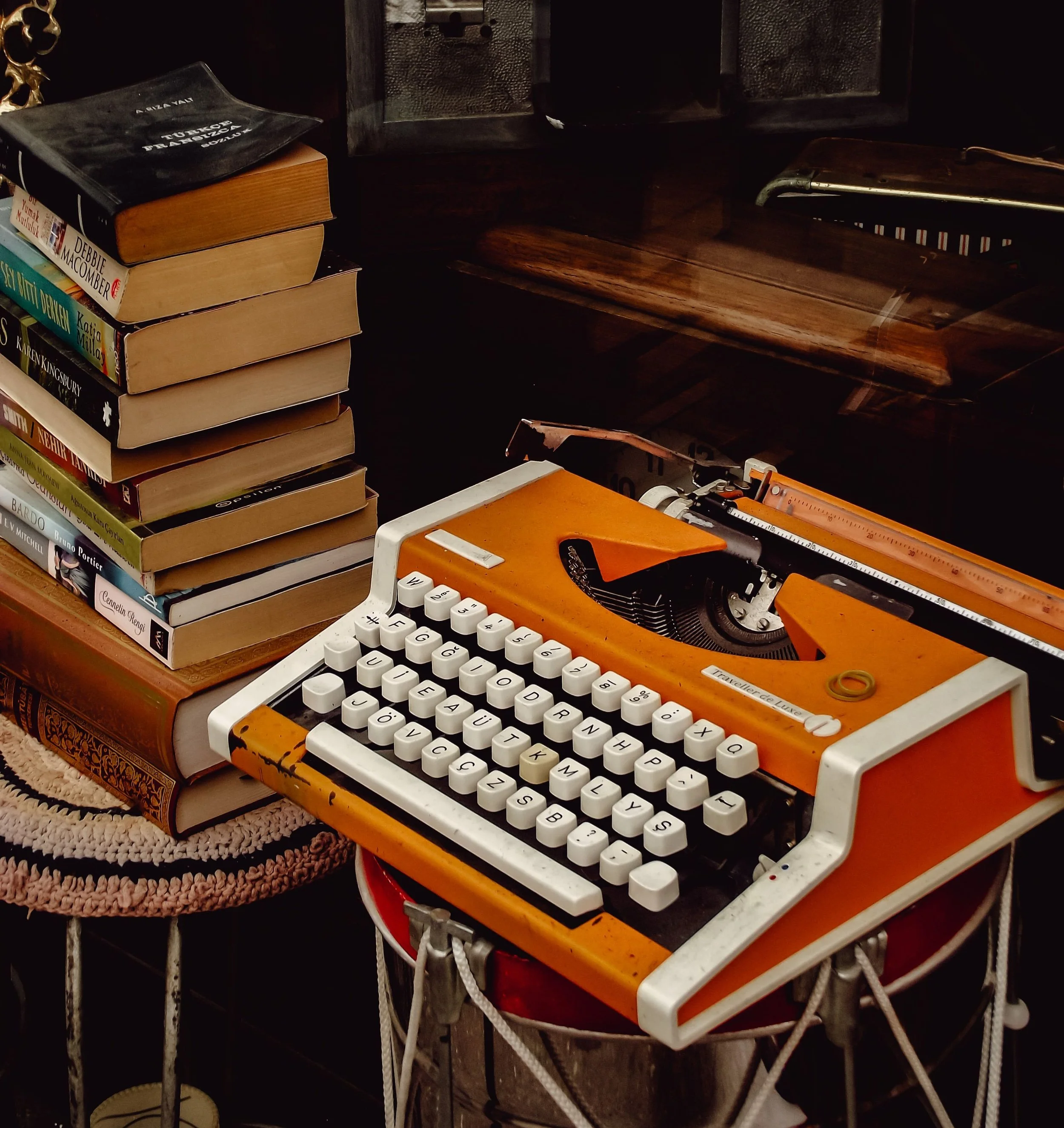 A vintage orange typewriter with white keys, sitting on a table next to a stack of books and a small round woven rug.