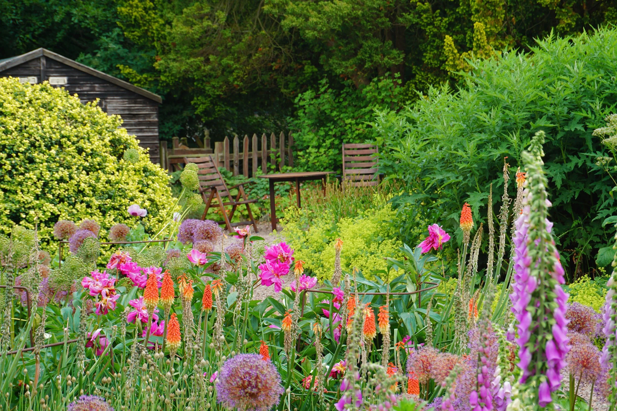 A lush garden scene with various colorful flowers in the foreground, a wooden shed, and a small seating area with chairs and a table surrounded by dense green trees in the background.