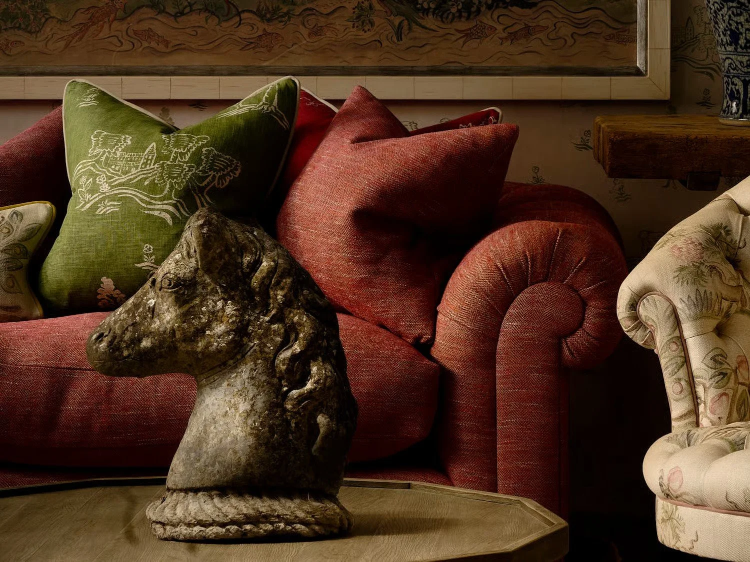 A decorative living room corner with a red upholstered sofa, various patterned pillows including green with white embroidery and floral, a beige floral armchair, a wooden side table, a framed artwork above the sofa, and a horse head sculpture on a wooden table in the foreground.