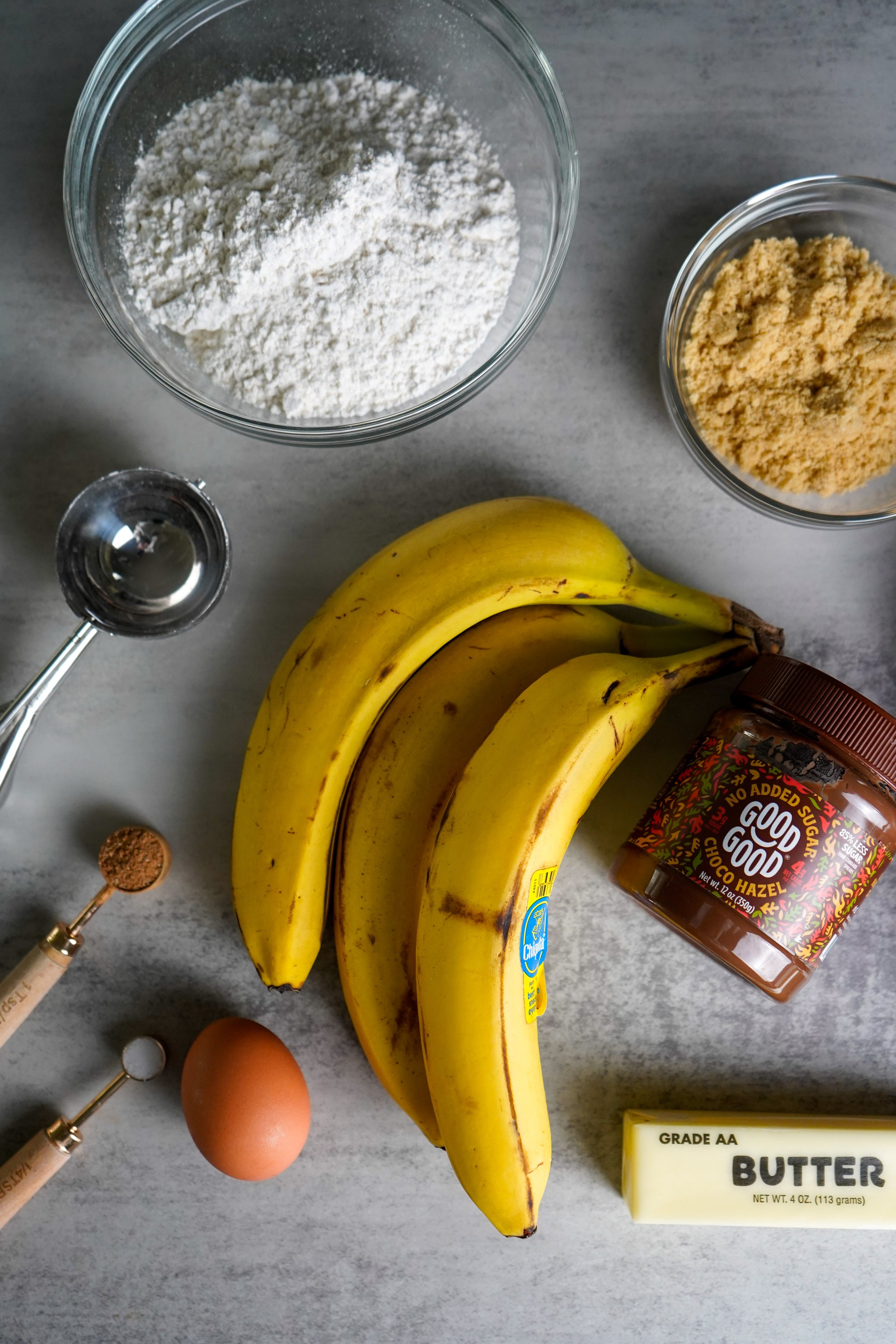 Bananas, flour, brown sugar, butter, egg, baking soda, brown sugar, measuring spoons, and a jar of hazelnut spread.