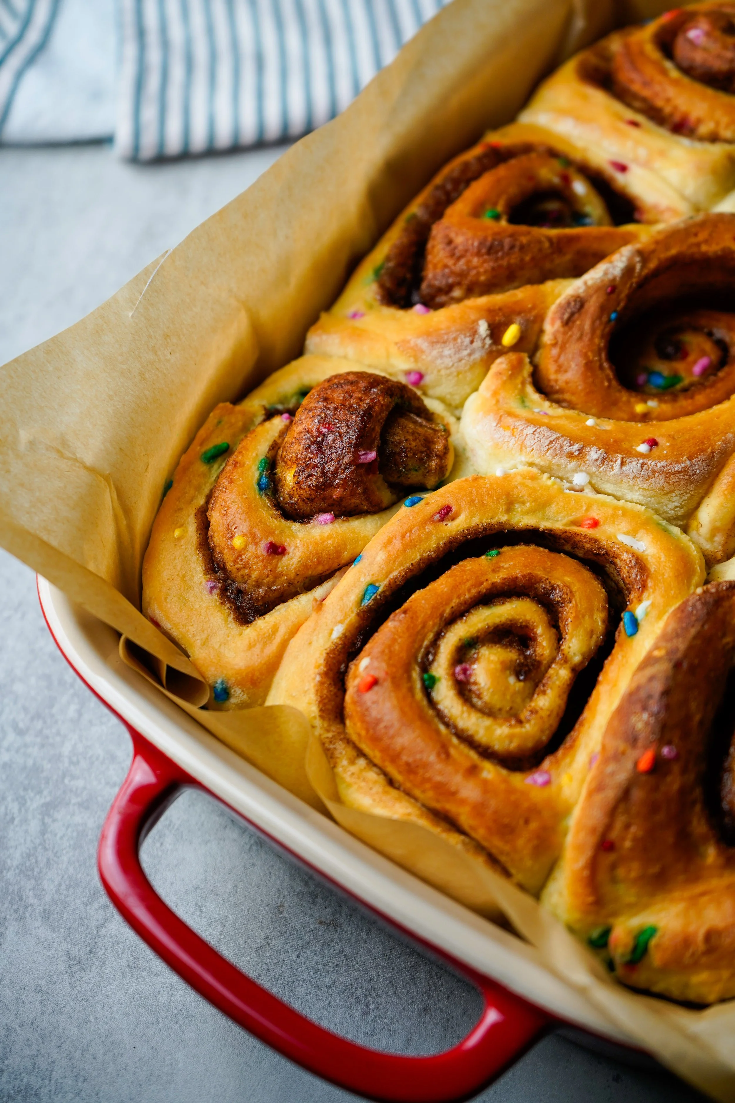 Baked cinnamon rolls with colorful sprinkles in a red baking dish lined with parchment paper.