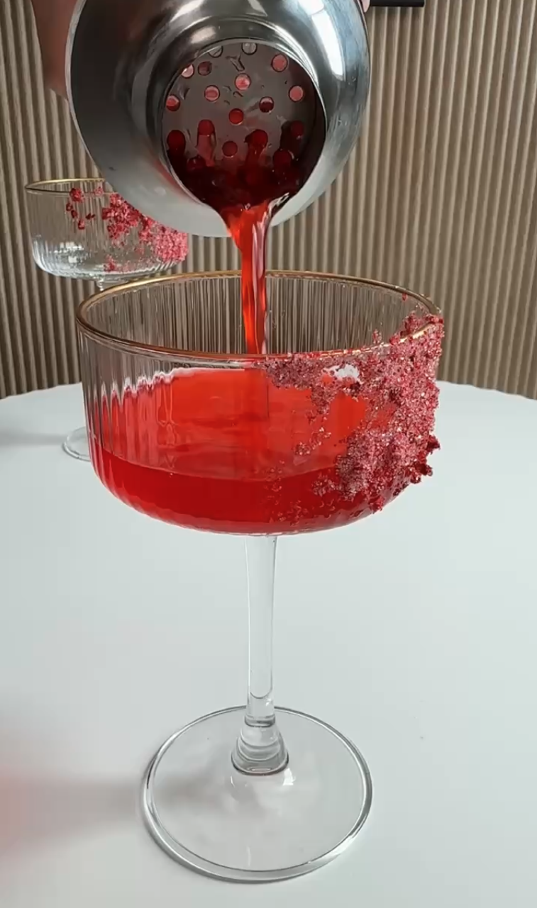 A red cocktail being poured from a shaker into a glass with a sugared rim, with another cocktail glass in the background on a white surface against a wooden panel wall.