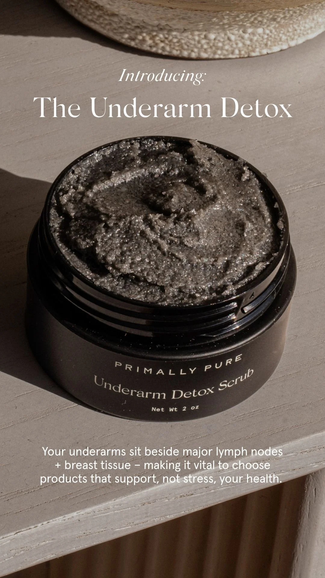 A black jar of Primally Pure Underarm Detox Scrub with a textured, grayish product inside, placed on a wooden surface.