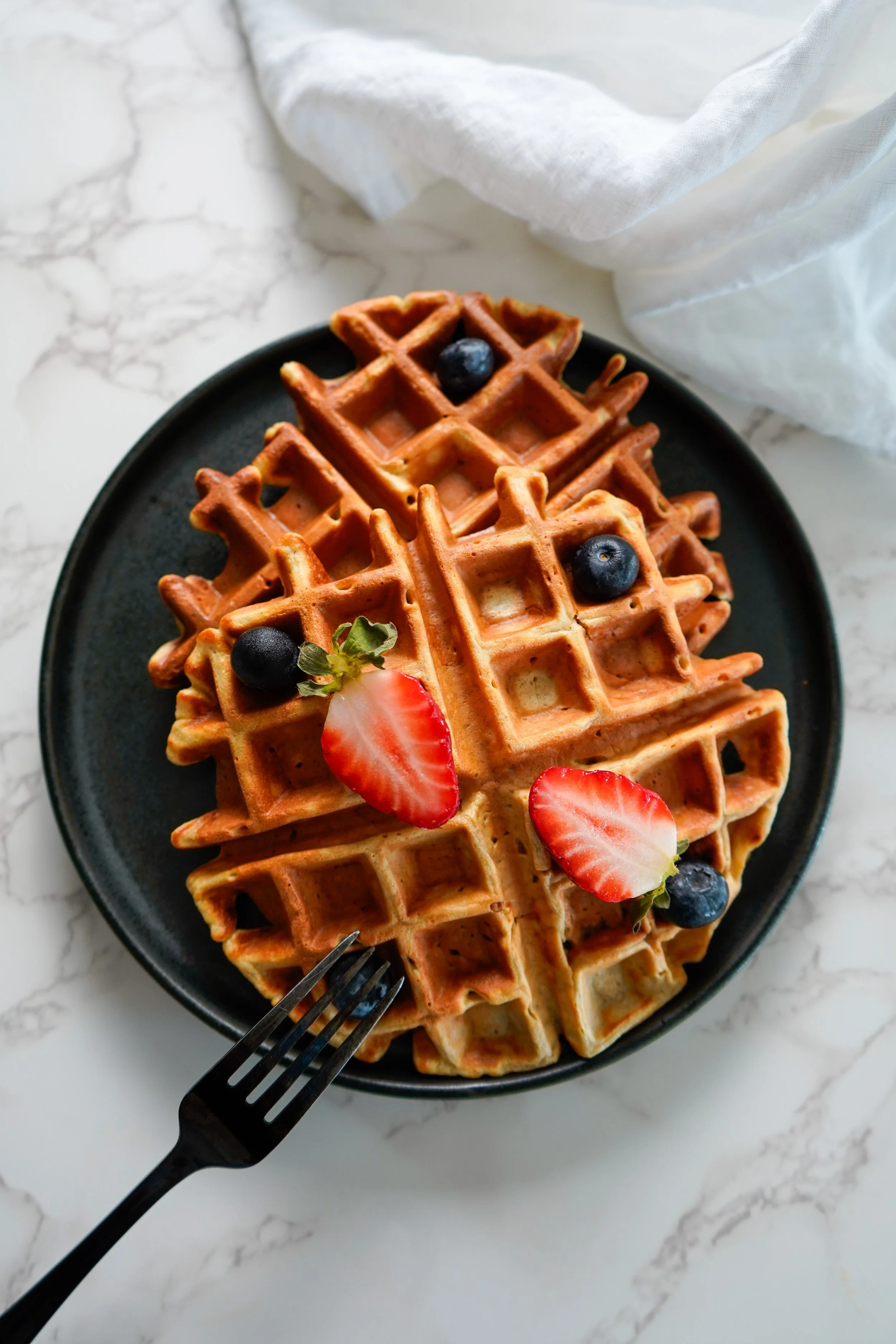 Two waffles topped with sliced strawberries and blueberries on a black plate, with a fork.