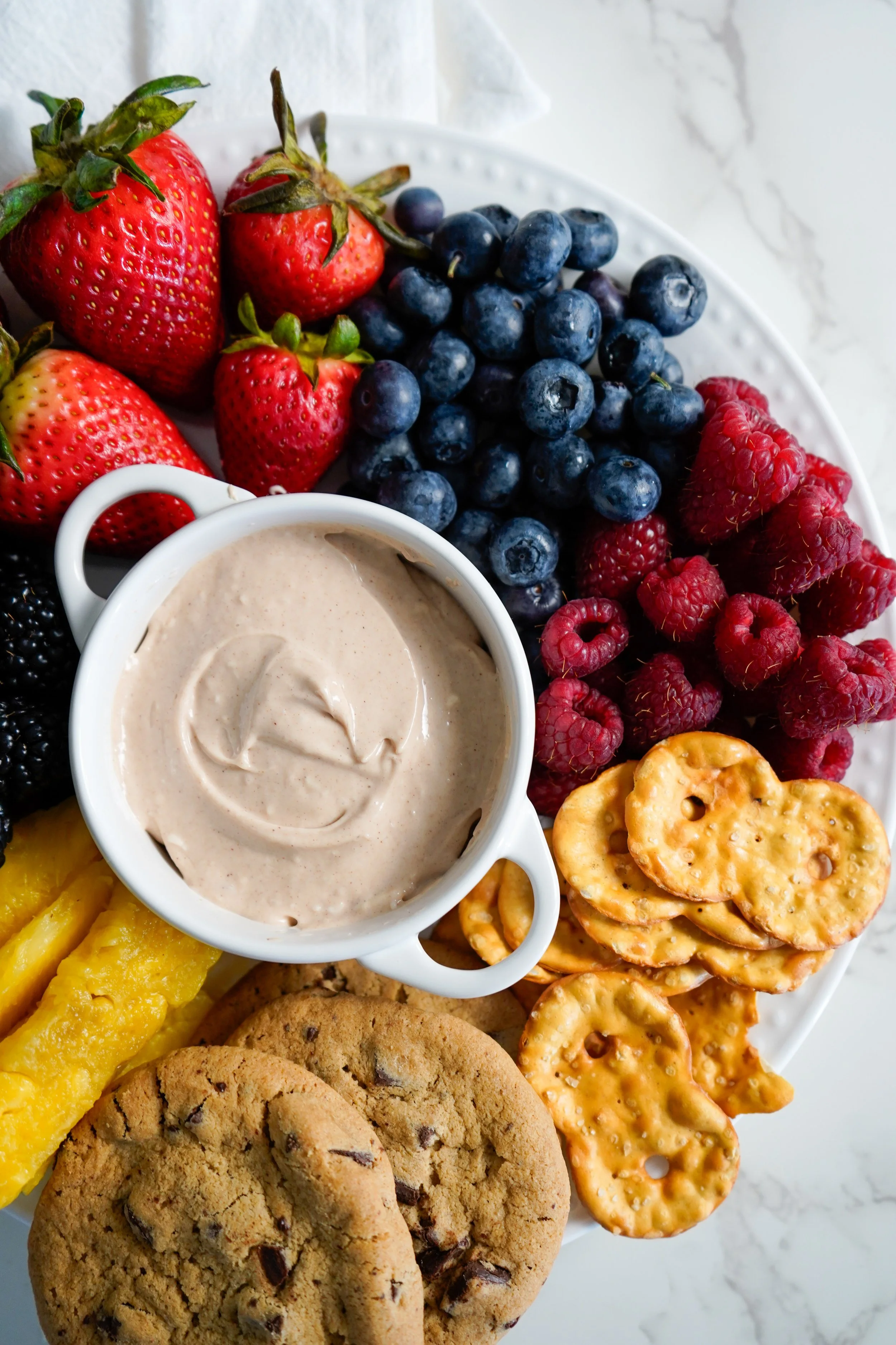A platter of fresh strawberries, blueberries, raspberries, blackberries, pineapple slices, cookies, pretzels, and a bowl of creamy dip on a white plate on a marble surface.