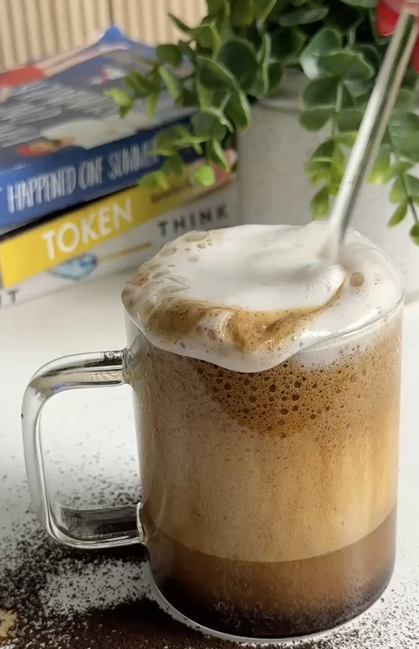 A glass mug of frothy coffee with foam and a straw, sprinkled with maple cinnamon, on a white surface with scattered ground coffee, with books and green plants in the background.