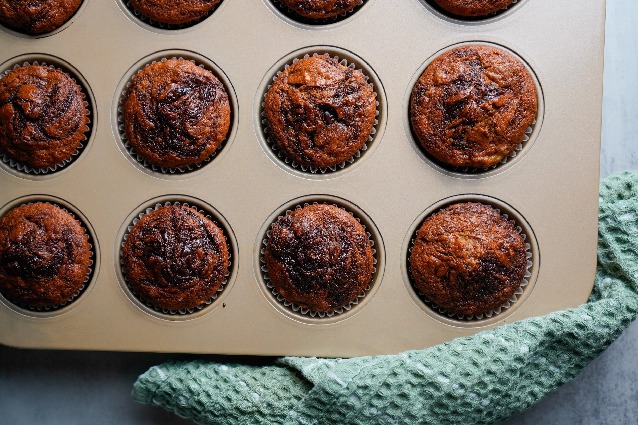 Top view of a muffin tin with nine banana muffins, some with darker or lighter spots, on a green textured cloth.