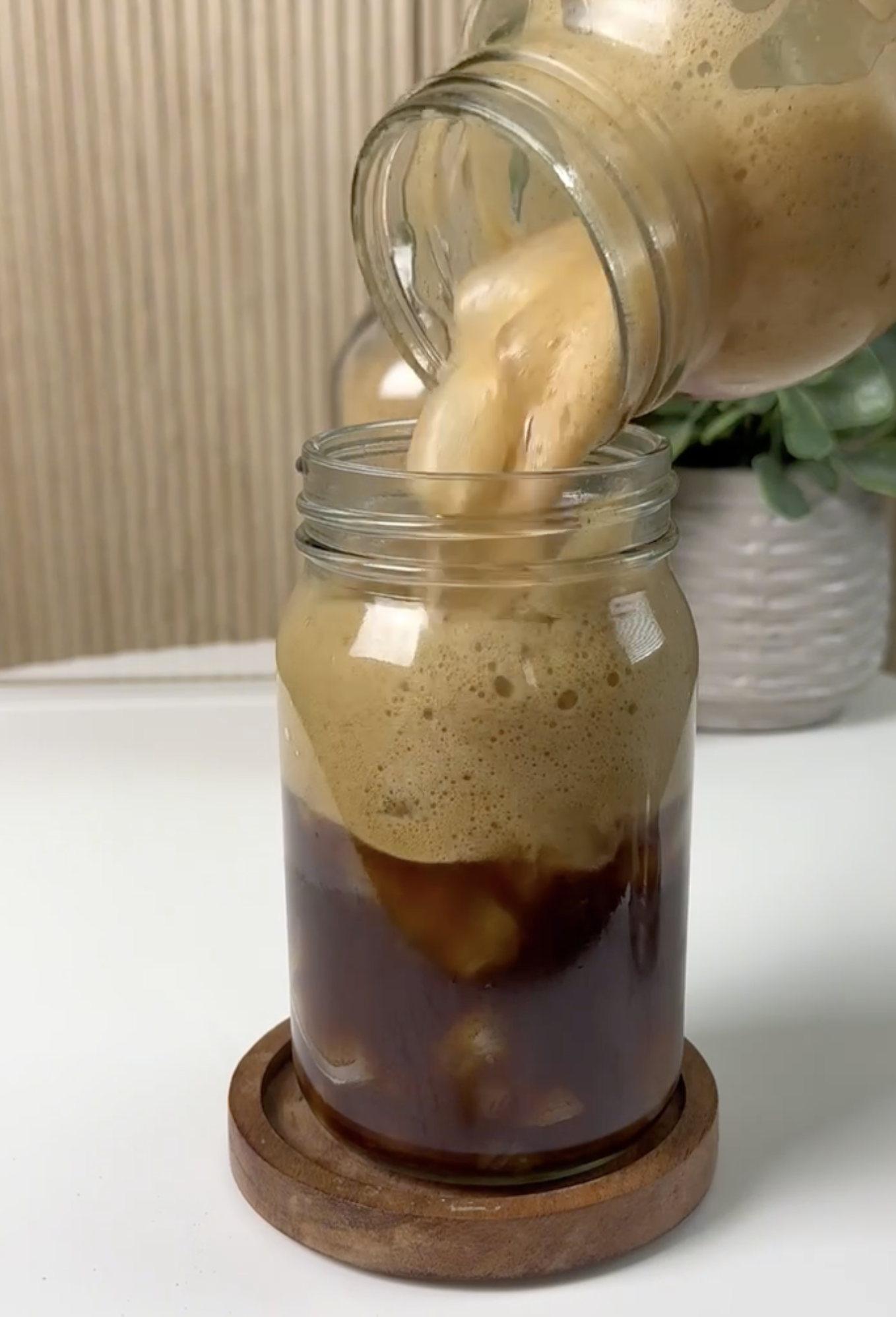 Pouring a iced coffee from a jar into a glass jar, with foam on top, on a wooden coaster.