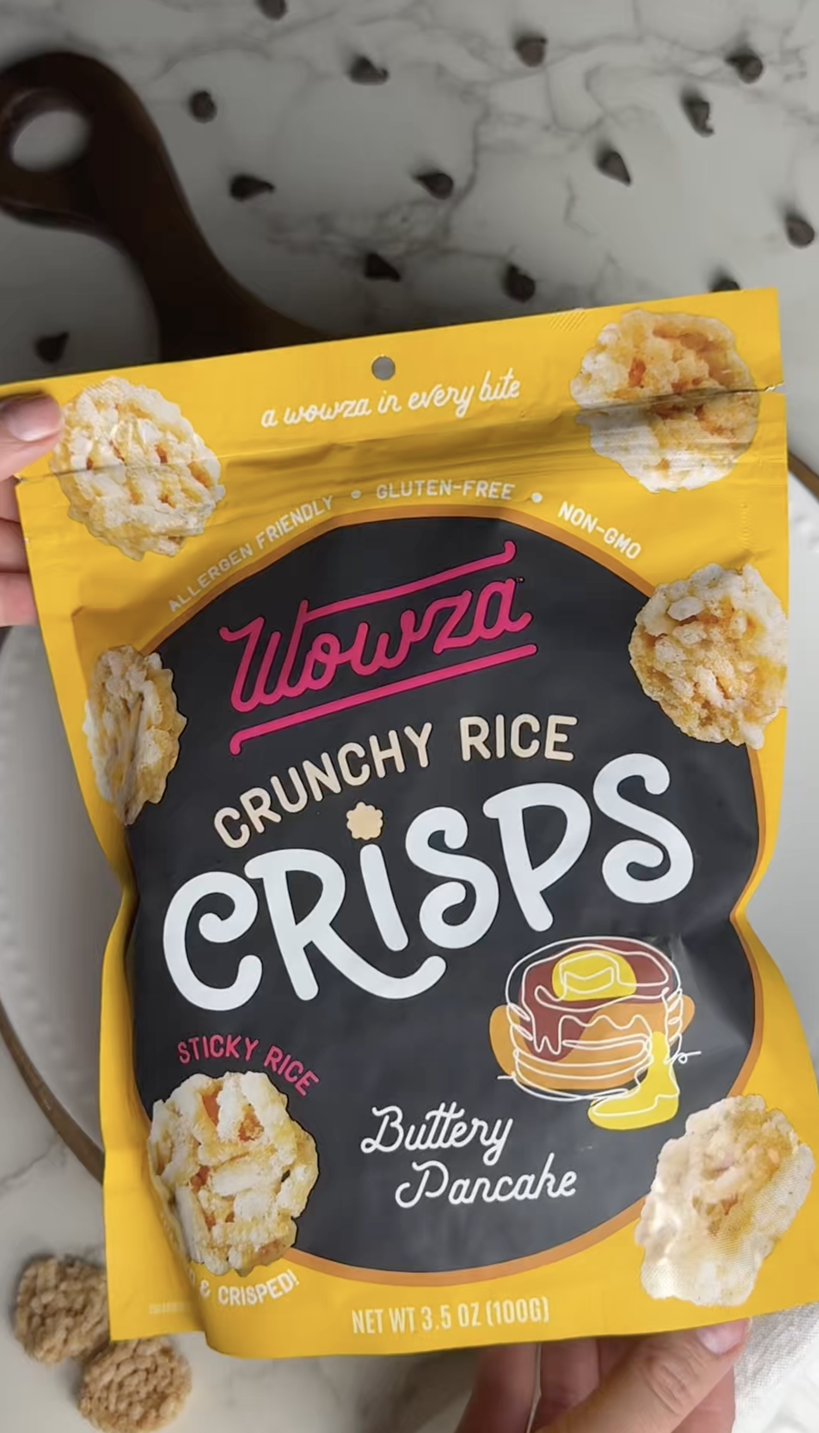 Packet of Wowza Crunchy Rice Crisps Buttery Pancake flavor rice cakes, package showing images of crispy rice snacks, with some snacks visible on a nearby plate and surface.
