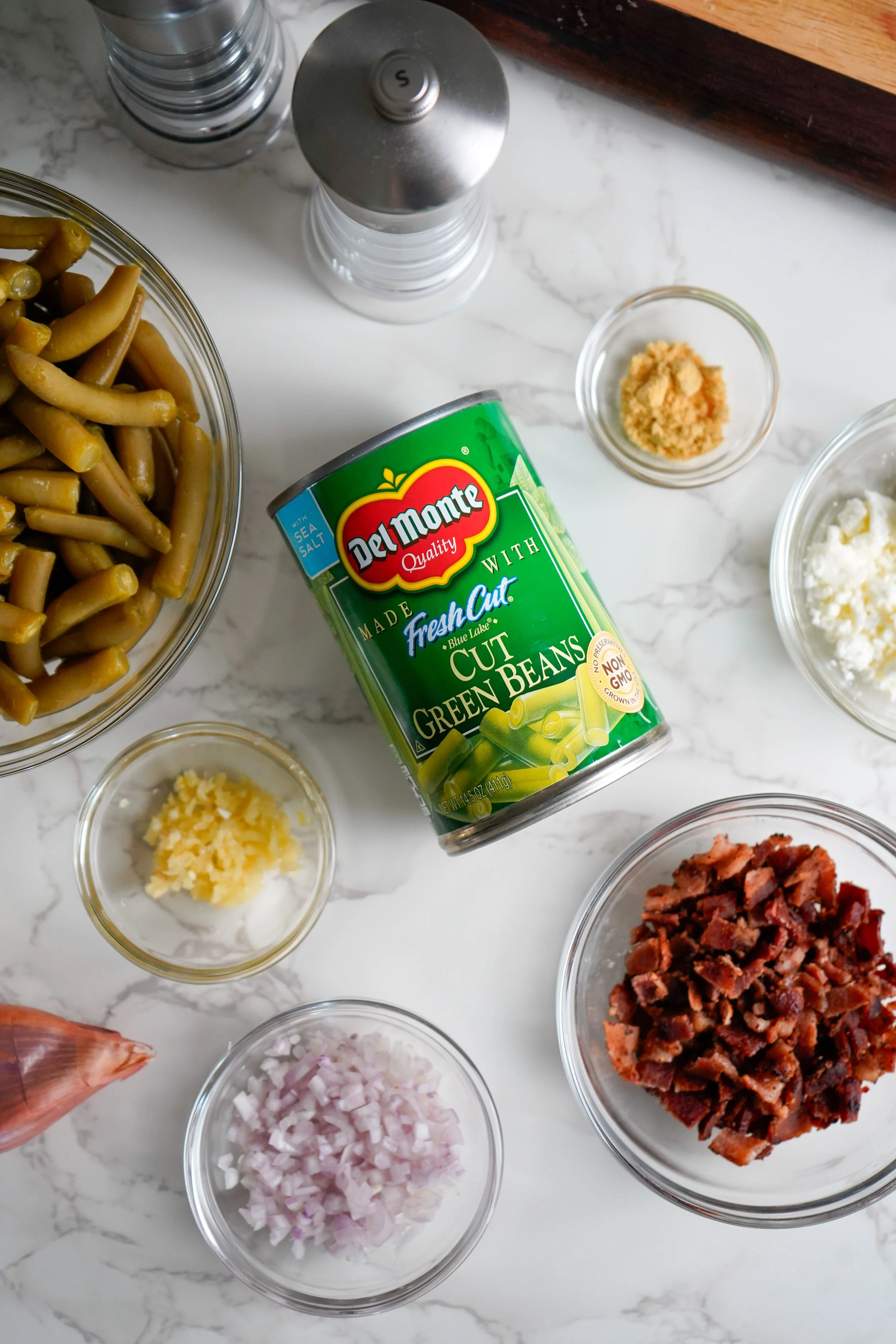 Canned cut green beans, chopped onions, cooked bacon bits, minced garlic, crumbled feta cheese, salt, and pepper on a marble countertop, with salt and pepper shakers in the background.