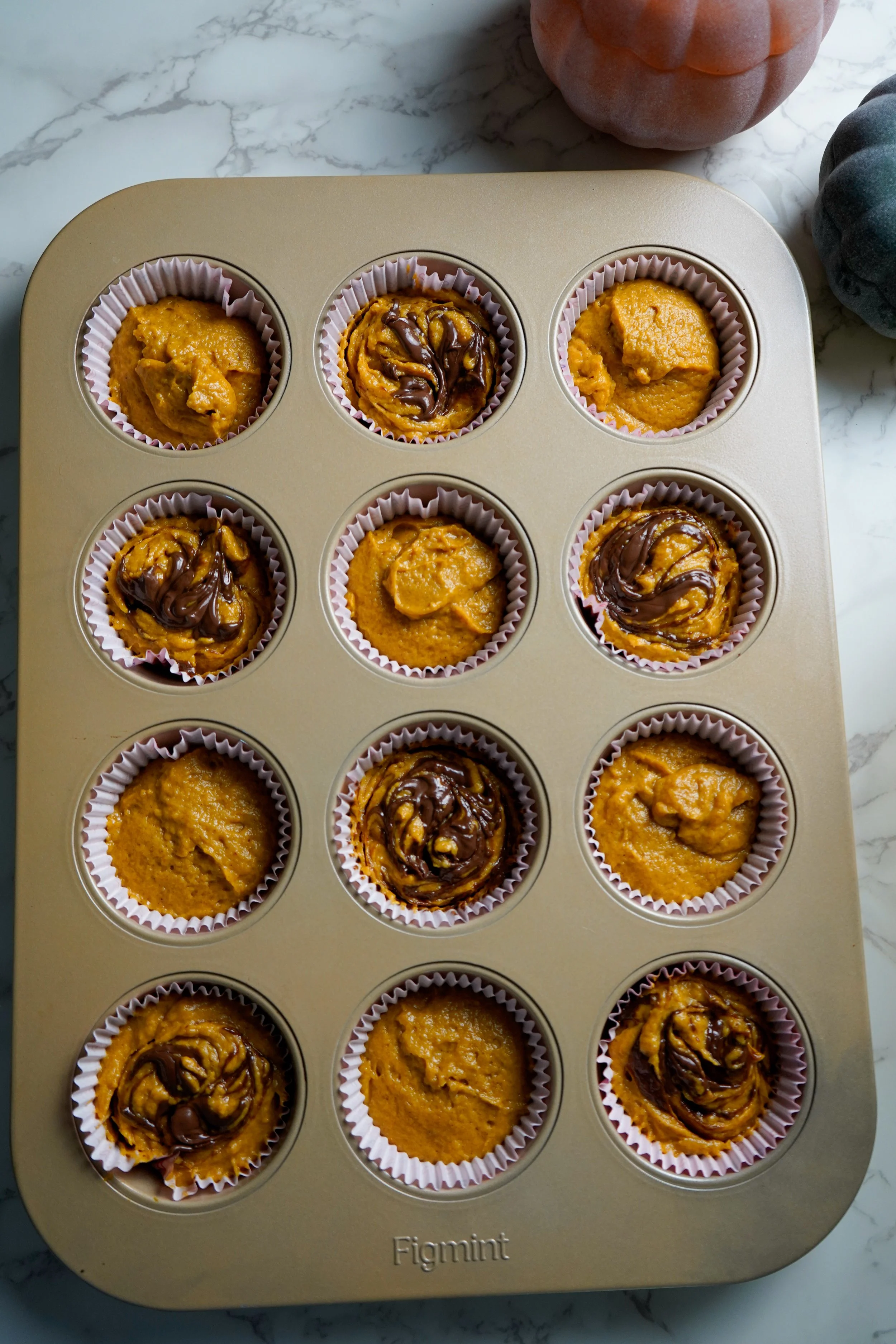 A baking pan filled with twelve cupcake liners, each containing pumpkin muffin batter, some topped with hazelnut swirls, on a marble countertop.