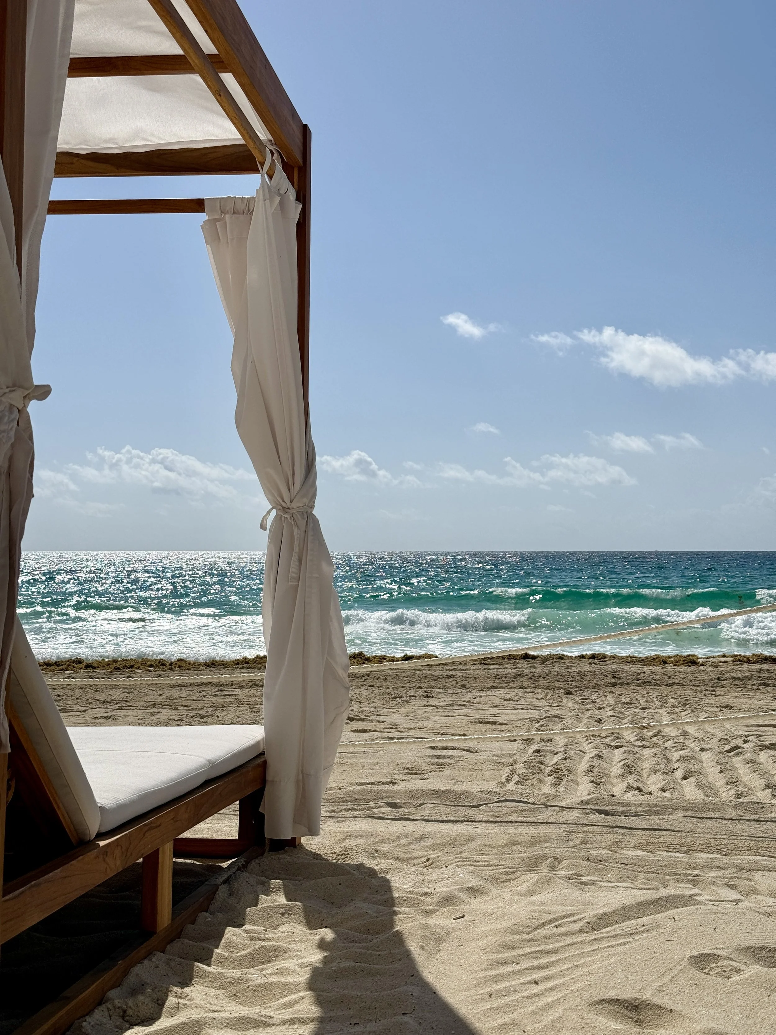 A beach scene with a wooden cabana and white curtains, sandy shore, and the ocean with blue waves and a clear sky with a few clouds.