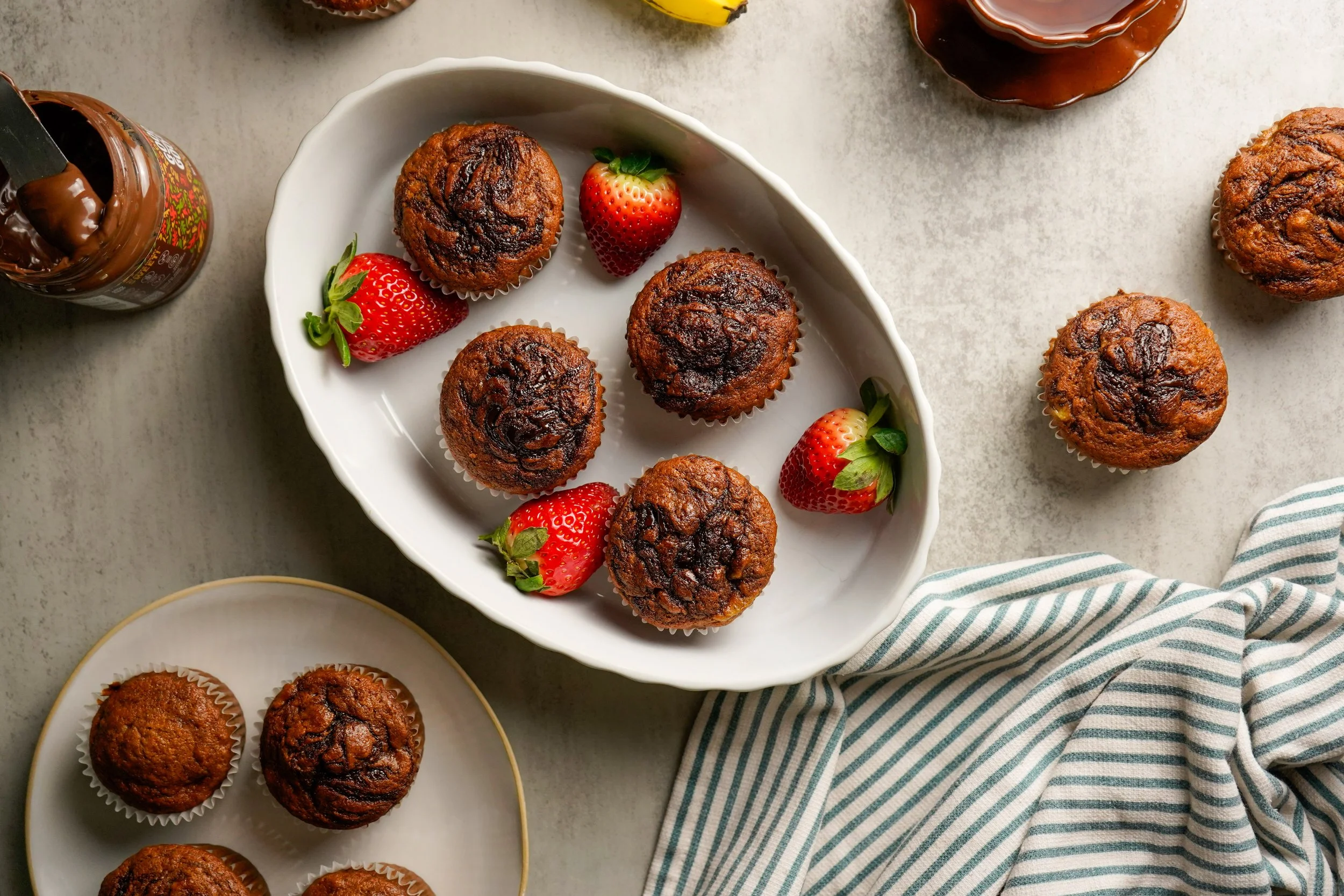 Hazelnut swirl banana muffins with strawberries on a white serving dish, with additional muffins nearby and a striped cloth on a light surface.