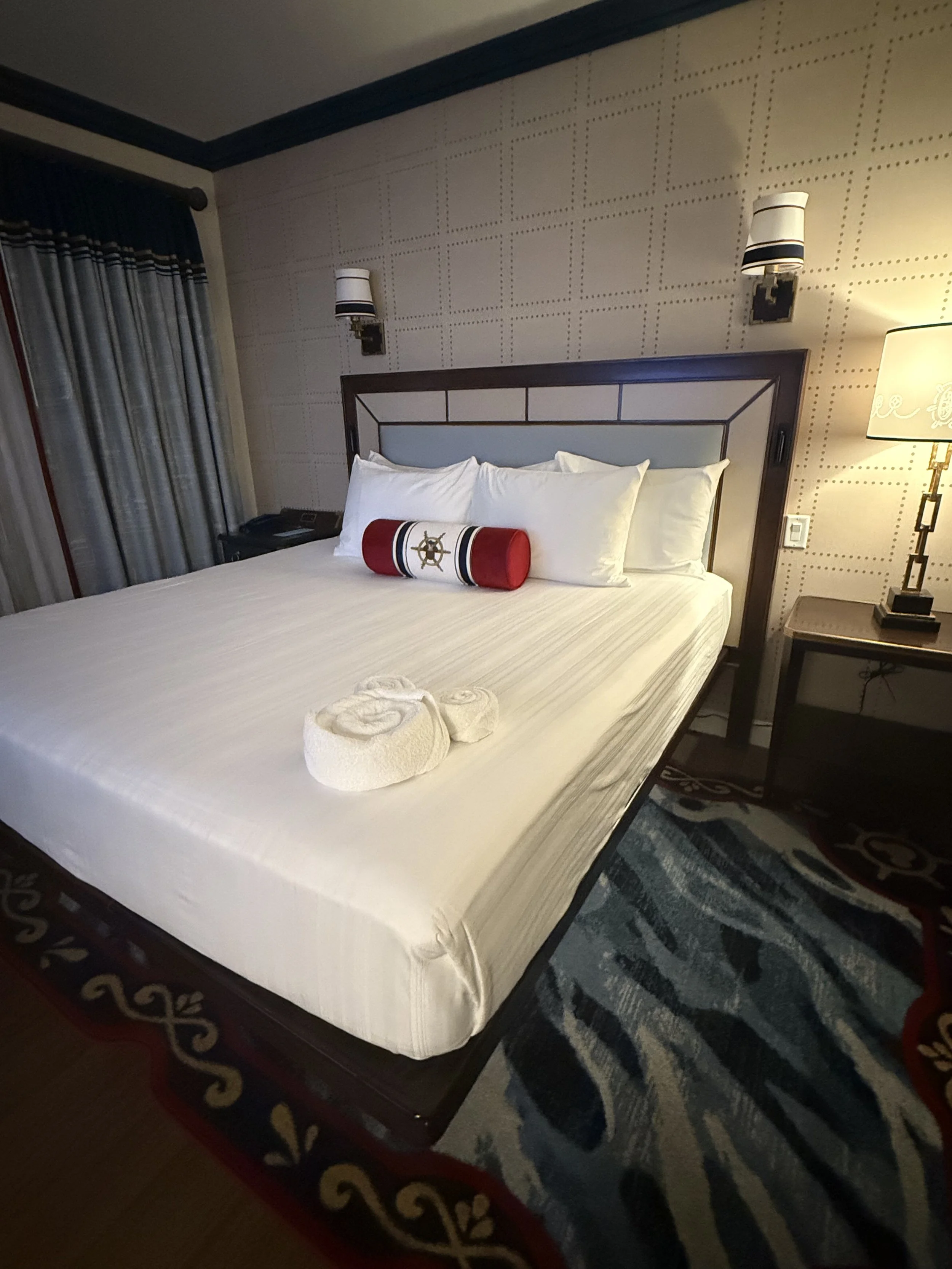 A neatly made hotel bed with white sheets and pillows, decorated with a red and white cylindrical pillow with a ship's wheel emblem, and set in a room with patterned wallpaper, a side table with a lamp, and a window with curtains.