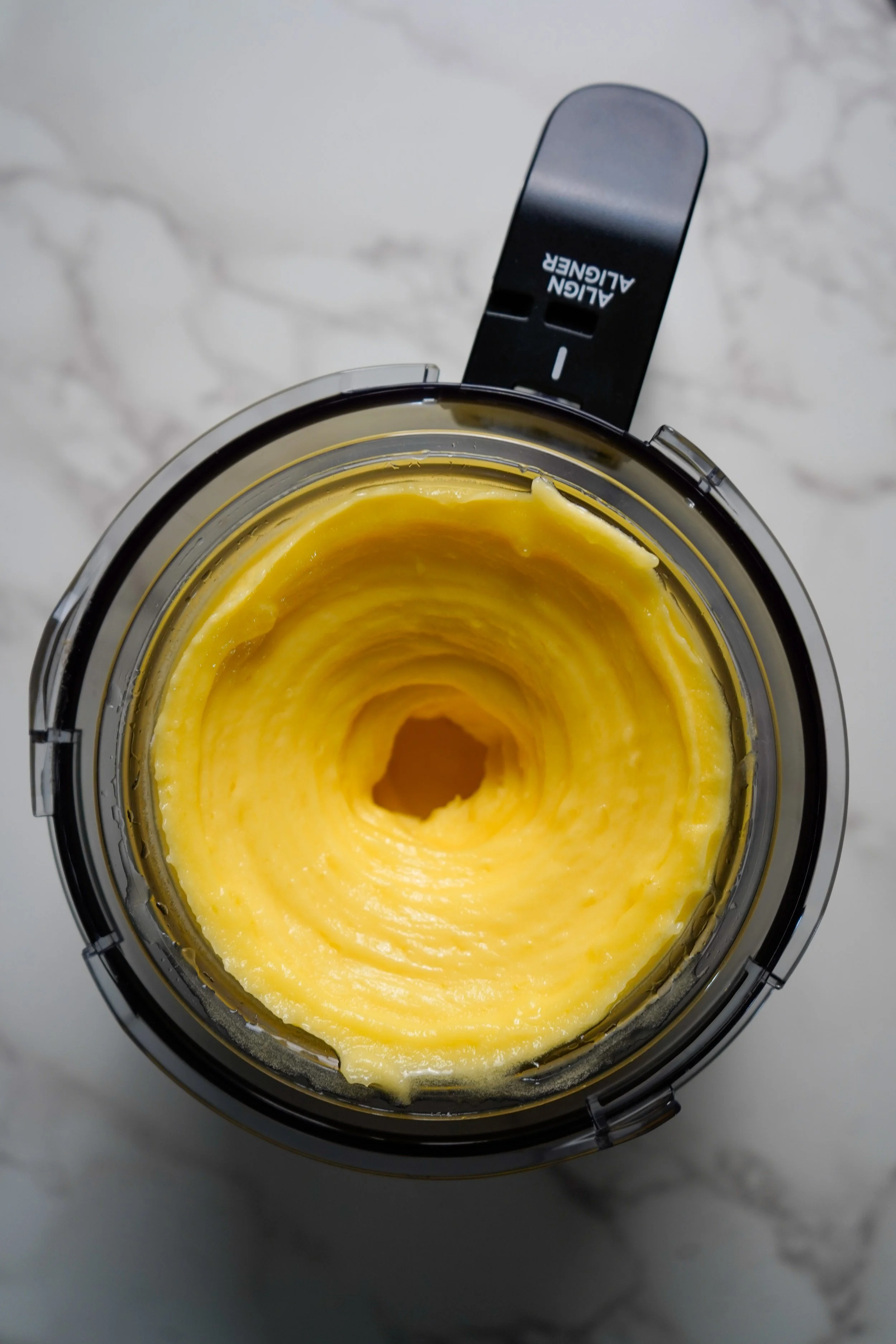 Inside a Ninja Creami blender containing smooth, yellow, creamy sorbet mixture, viewed from above, with a marble countertop background.