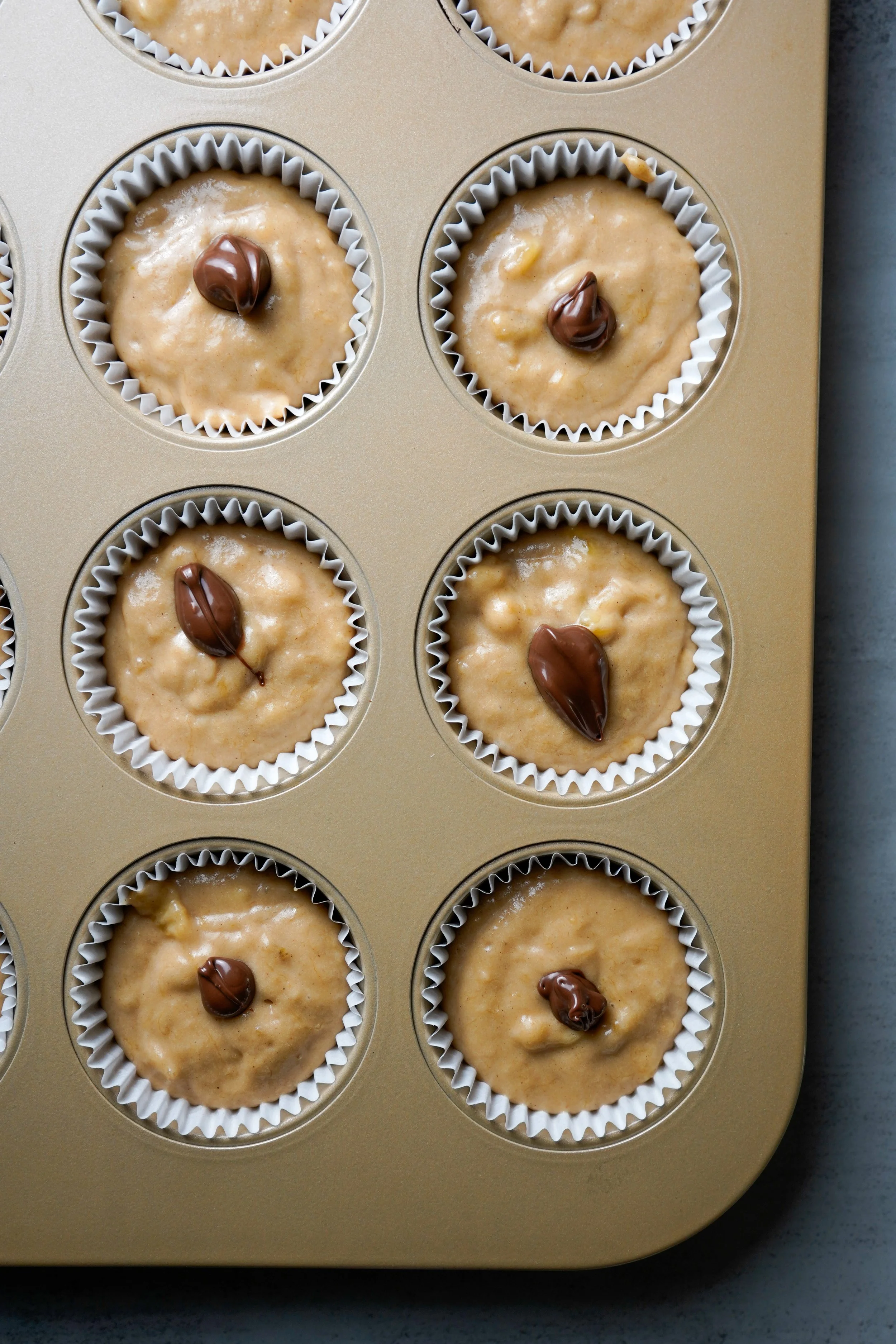 Batter-filled cupcake liners in a muffin pan, each topped with a small dollop of hazelnut spread.