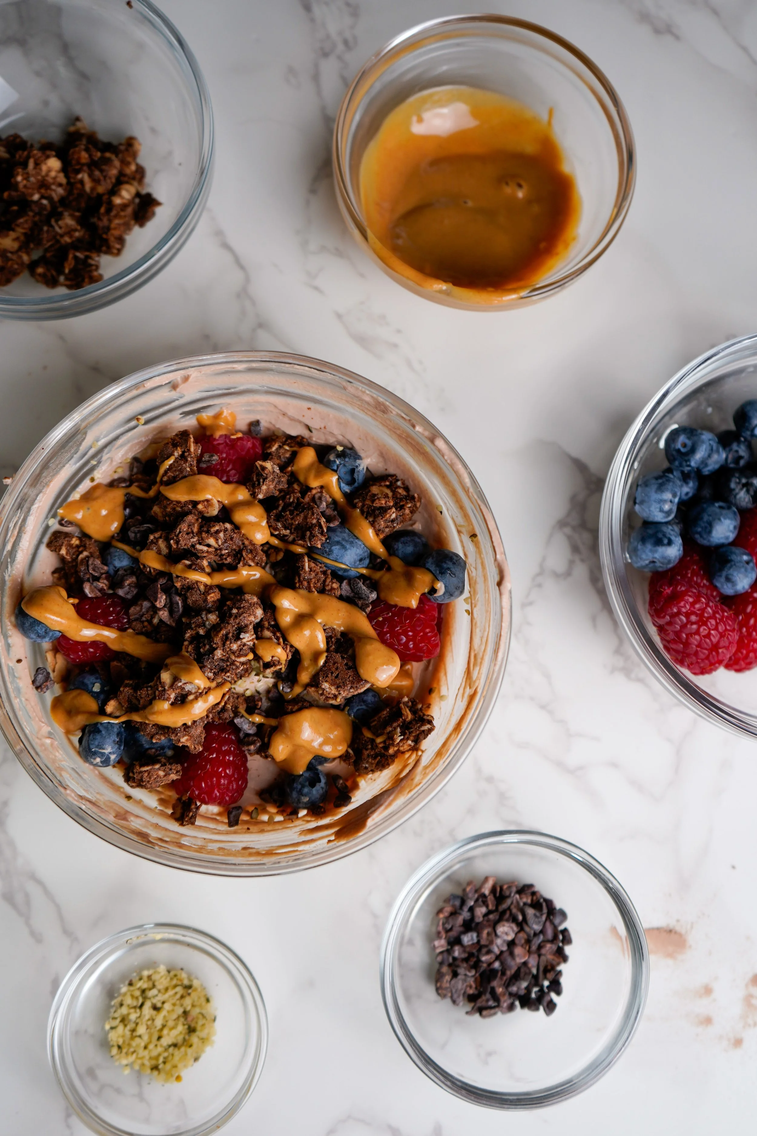 A protein yogurt bowl in a glass bowl topped with berries, chocolate, and peanut butter drizzle, with small bowls of ingredients surrounding it on a white marble surface.