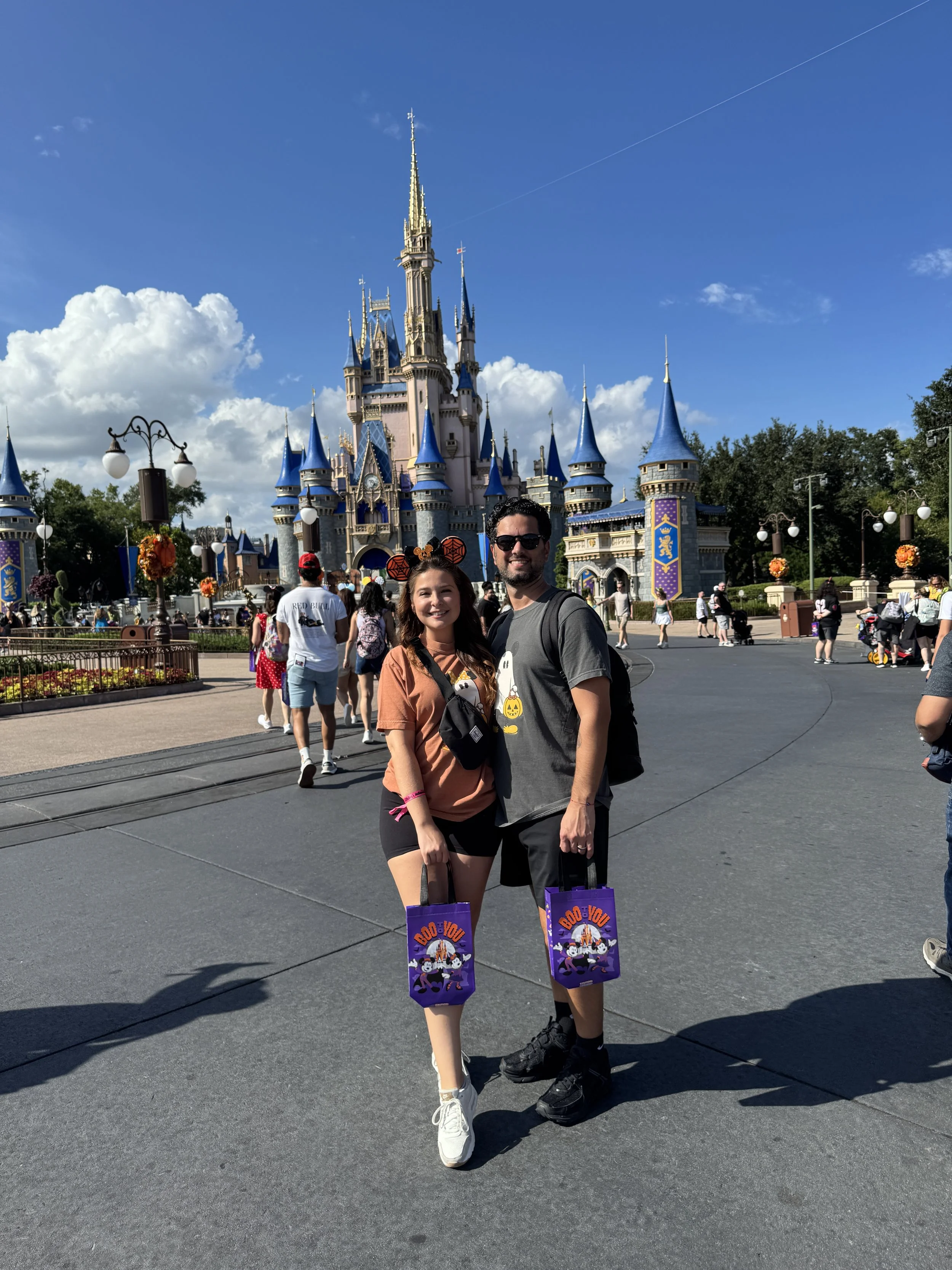 A happy couple standing in front of Cinderella Castle at Disneyland, holding "Boo You" trick-or-treat bags, with other visitors and the castle in the background on a sunny day.