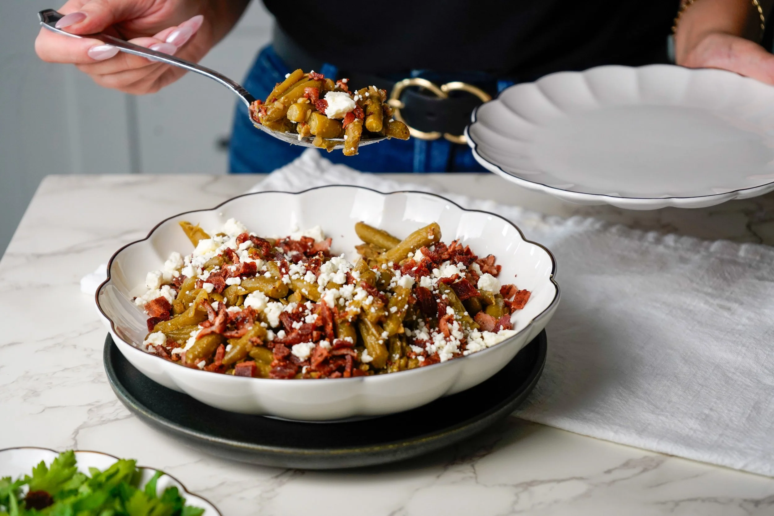 A person serving green beans topped with bacon bits and crumbled cheese into a white dish.