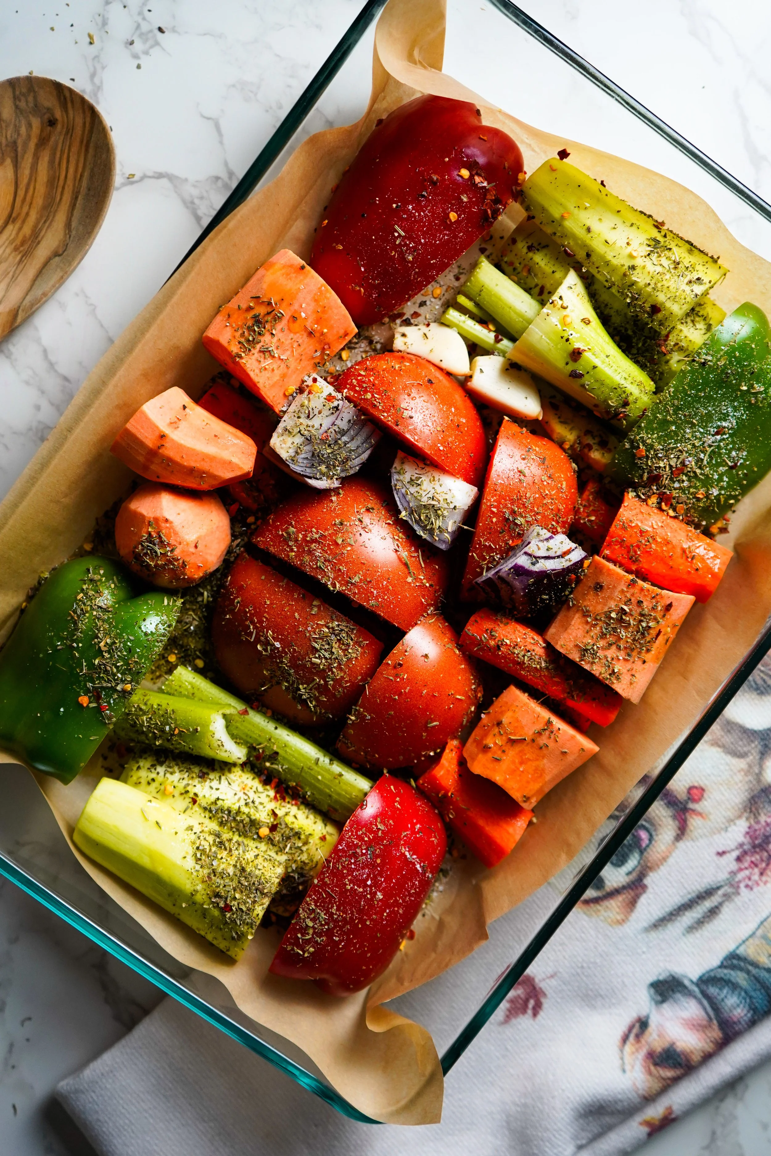 A glass baking dish filled with chopped vegetables including tomatoes, celery, and peppers, sprinkled with herbs and spices, on a marble surface with a wooden spoon and a cloth nearby.