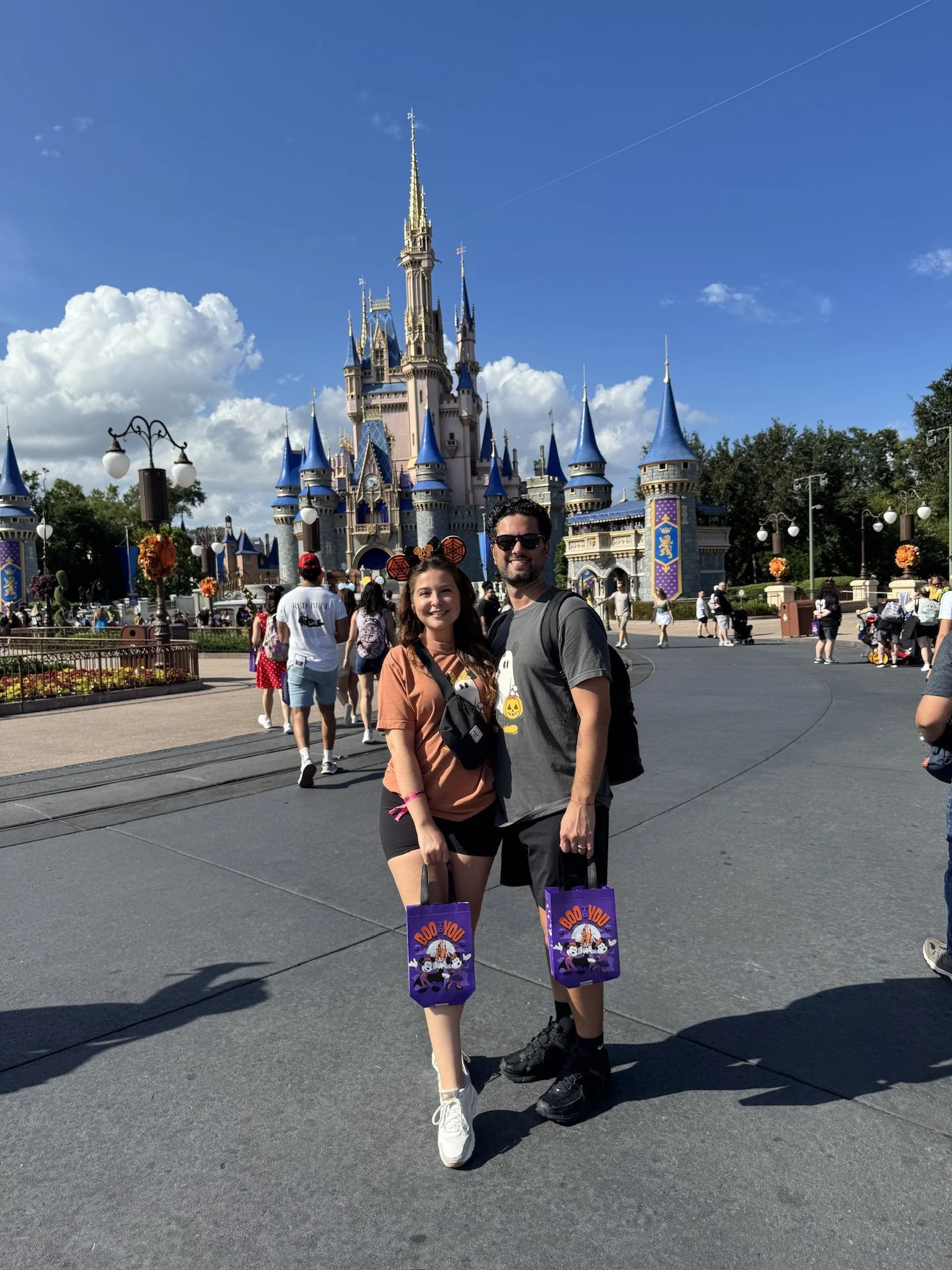 A couple standing in front of Sleeping Beauty Castle at Disneyland, holding Halloween-themed bags with Mickey Mouse and pumpkins against a sunny blue sky.
