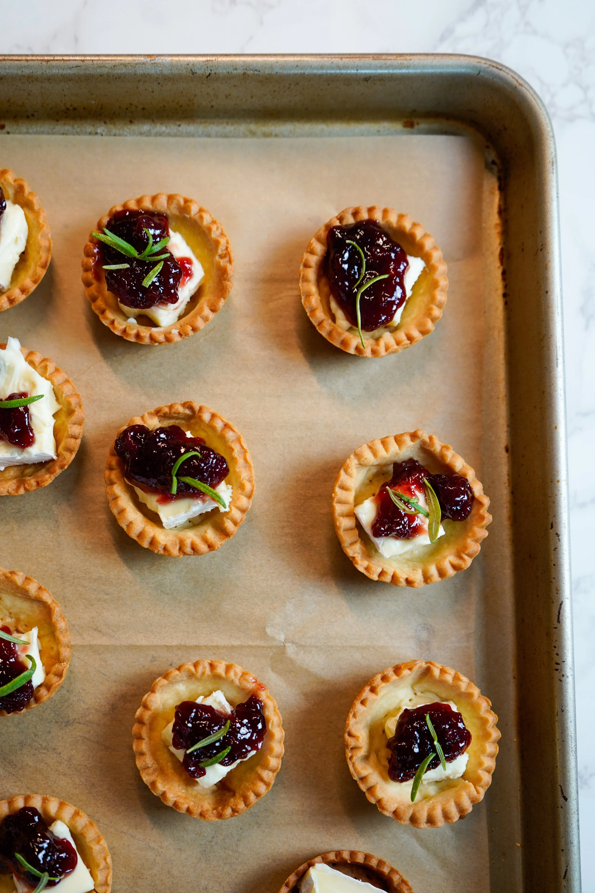 Mini tartlets topped with dollops of cheese, dark cherry spread, and garnished with small green herbs on a baking sheet lined with parchment paper.
