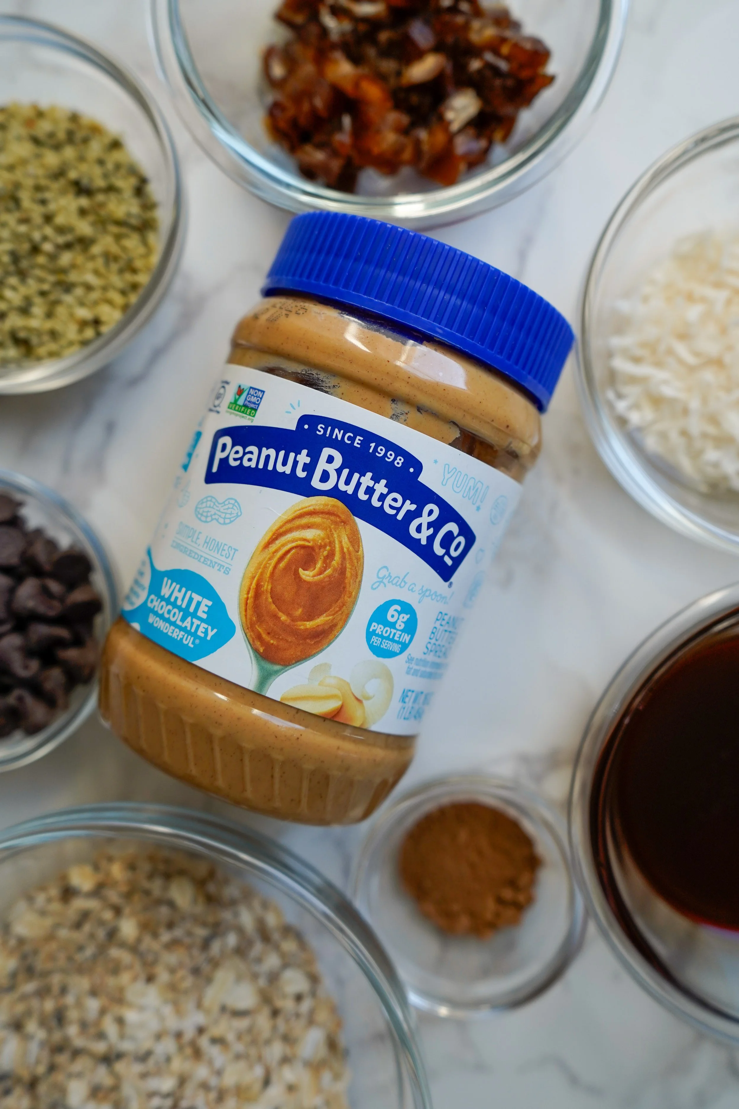 Jar of Peanut Butter & Co. white chocolate peanut butter surrounded by various ingredients in bowls, including chopped dates, shredded coconut, rolled oats, chocolate chips, hemp seeds, and cinnamon on a marble surface.