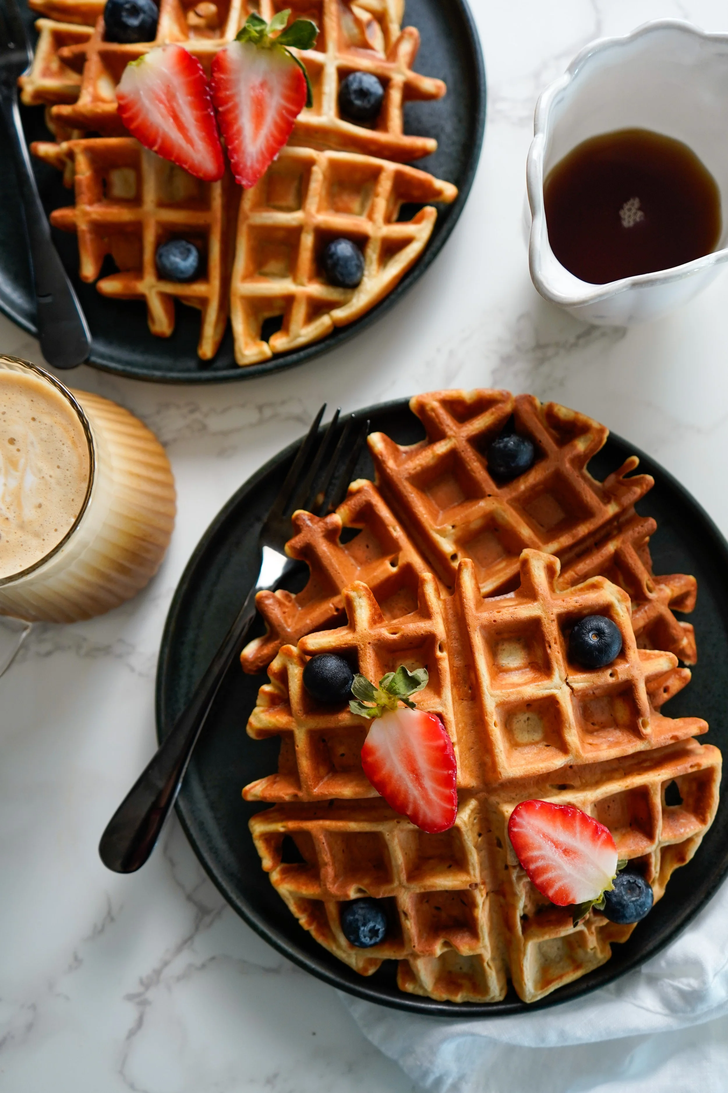 Two plates of waffles topped with strawberries and blueberries, accompanied by a cup of coffee and a glass of latte, on a white marble surface.
