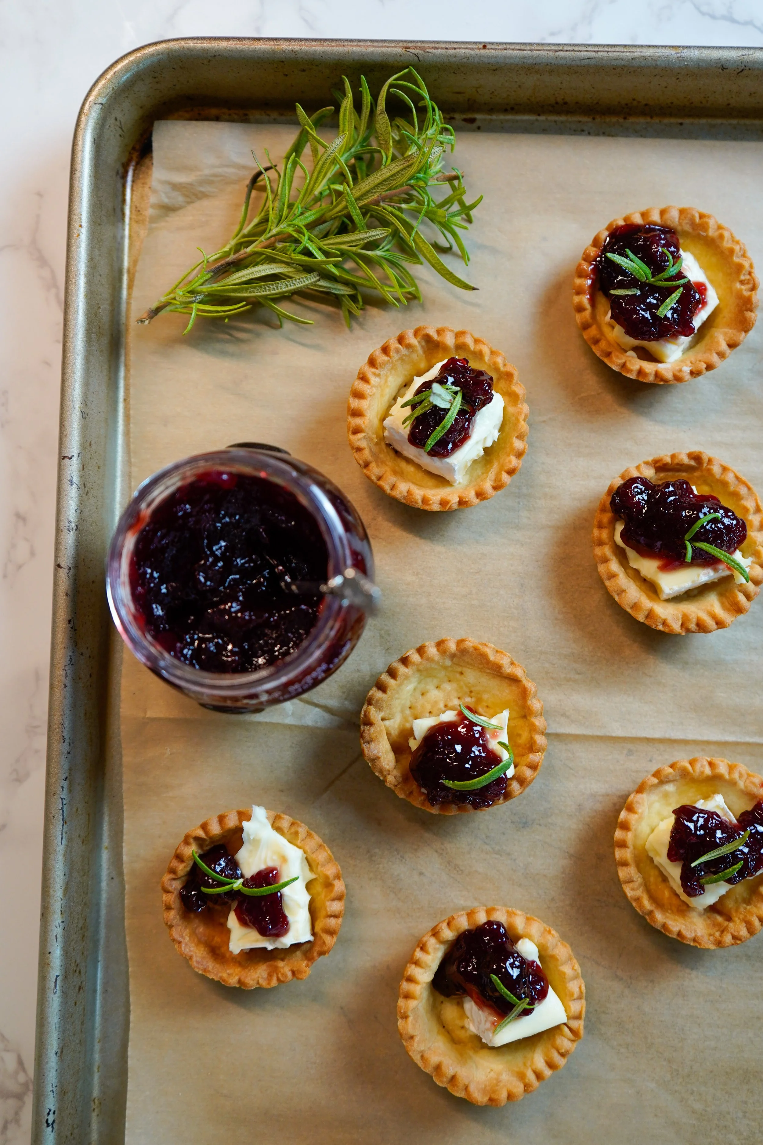 Mini tartlets topped with cheese, sour cherry jam, and a sprig of green herb, served on a baking sheet with a jar of jam and a sprig of fresh herbs.