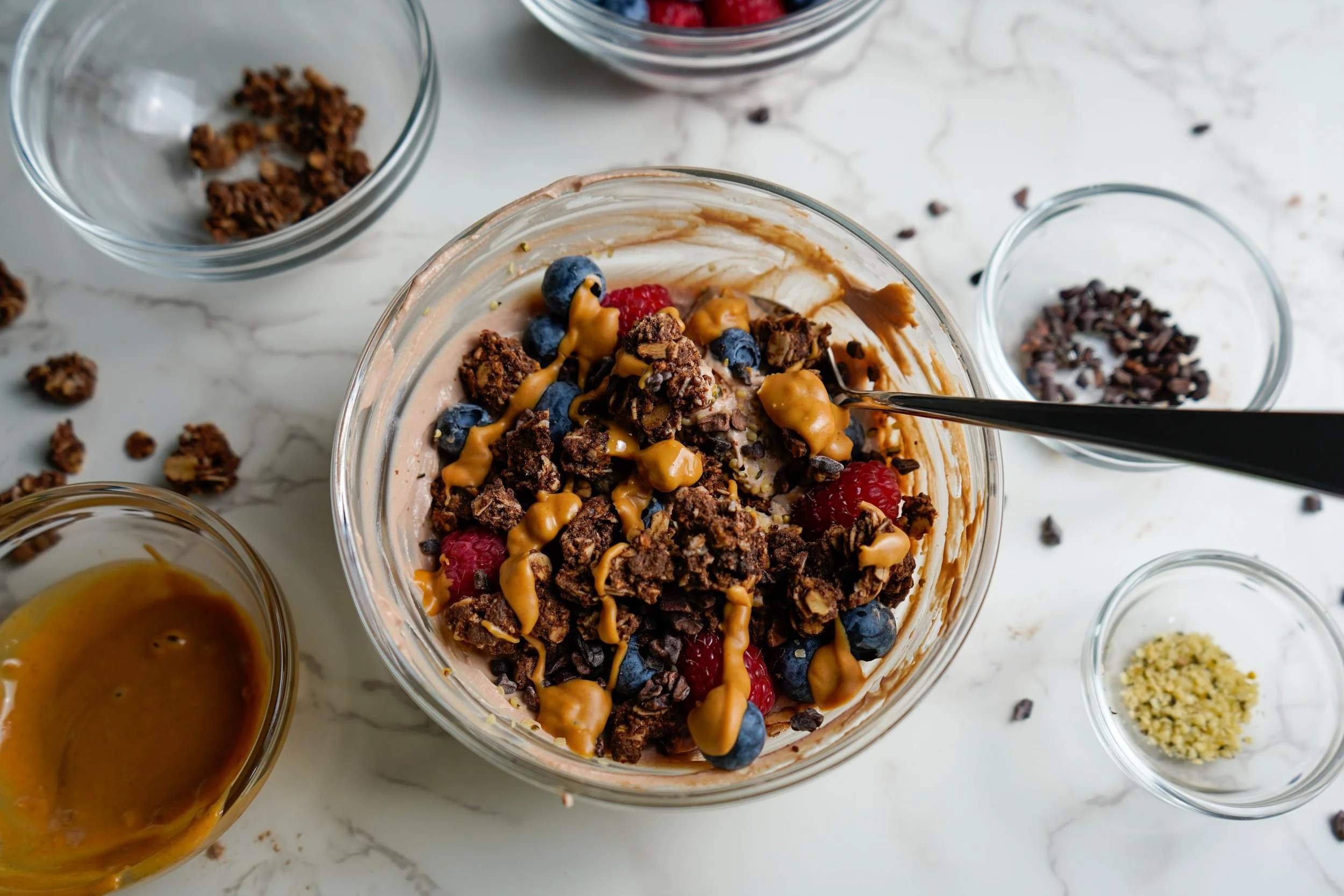 A glass bowl of protein yogurt topped with blueberries, raspberries, cacao nibs, peanut butter drizzle, and granola, surrounded by small bowls with more cacao nibs, peanut butter, and granola on a white marble surface.