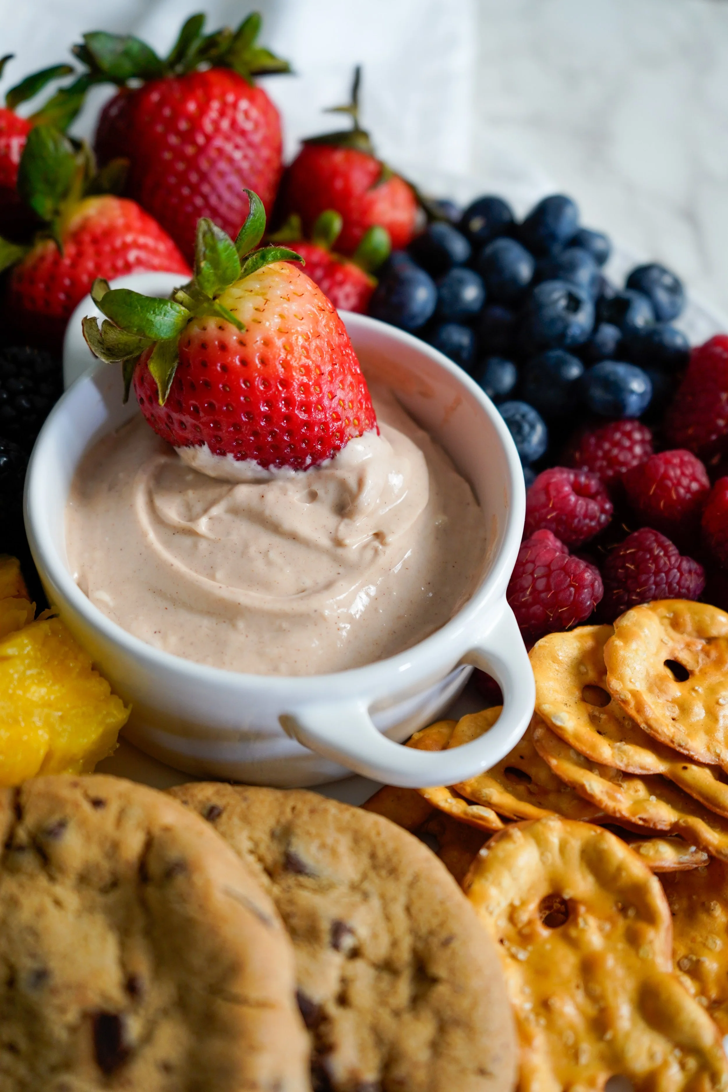 A bowl of churro fruit dip topped with a fresh strawberry surrounded by fresh strawberries, blueberries, raspberries, chocolate chip cookies, and pretzels.