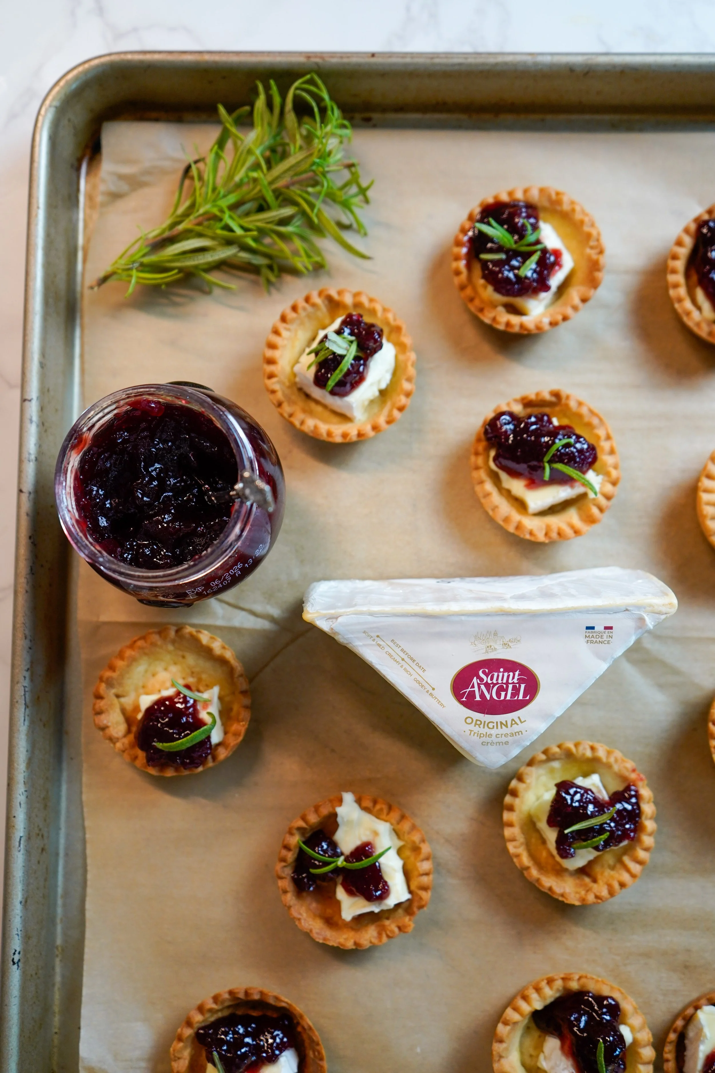 Mini tartlets topped with cheese and cherry jam, a wedge of Saint Angel triple cream cheese, a jar of sour cherry spread, and a sprig of fresh rosemary on a baking sheet lined with parchment paper.