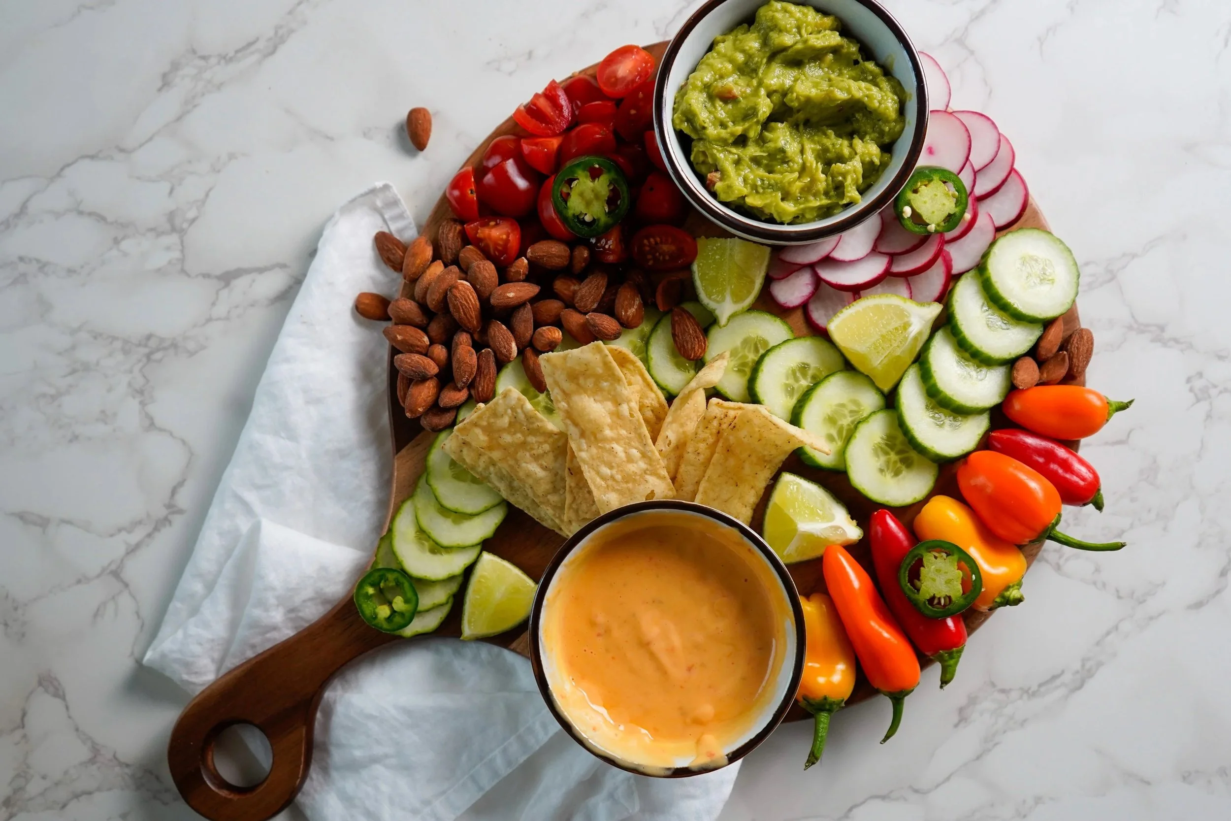 A charcuterie board with guacamole, cherry tomatoes, almonds, radishes, cucumbers, lime wedges, tortilla chips, a creamy dip, and assorted small peppers on a white marble surface.
