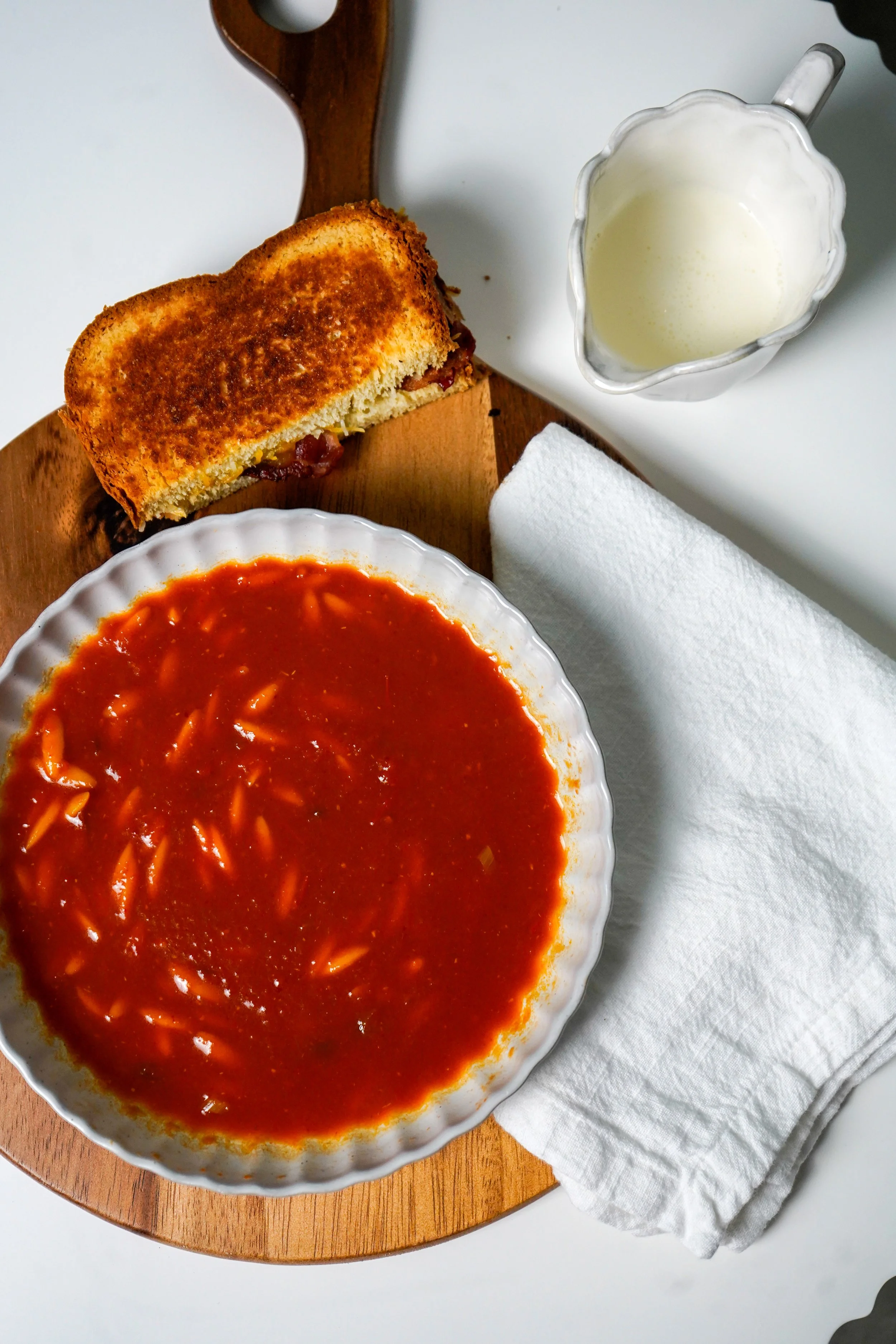 A bowl of tomato soup with pasta, a slice of grilled cheese sandwich, a glass of heavy cream, and a white napkin on a wooden tray.
