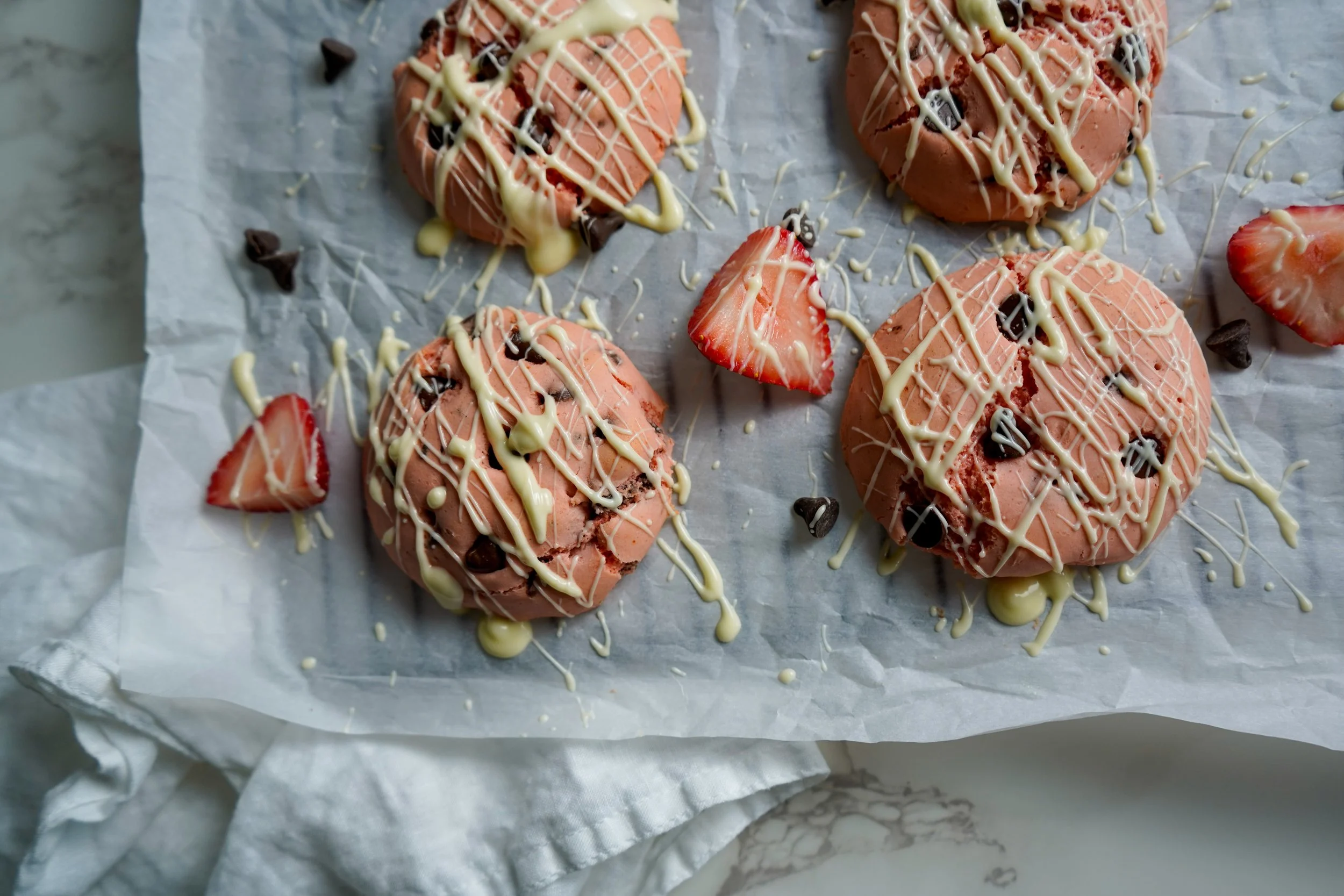 Four pink strawberry chocolate chip cookies topped with white chocolate drizzle, surrounded by sliced strawberries and small chocolate chips on parchment paper.