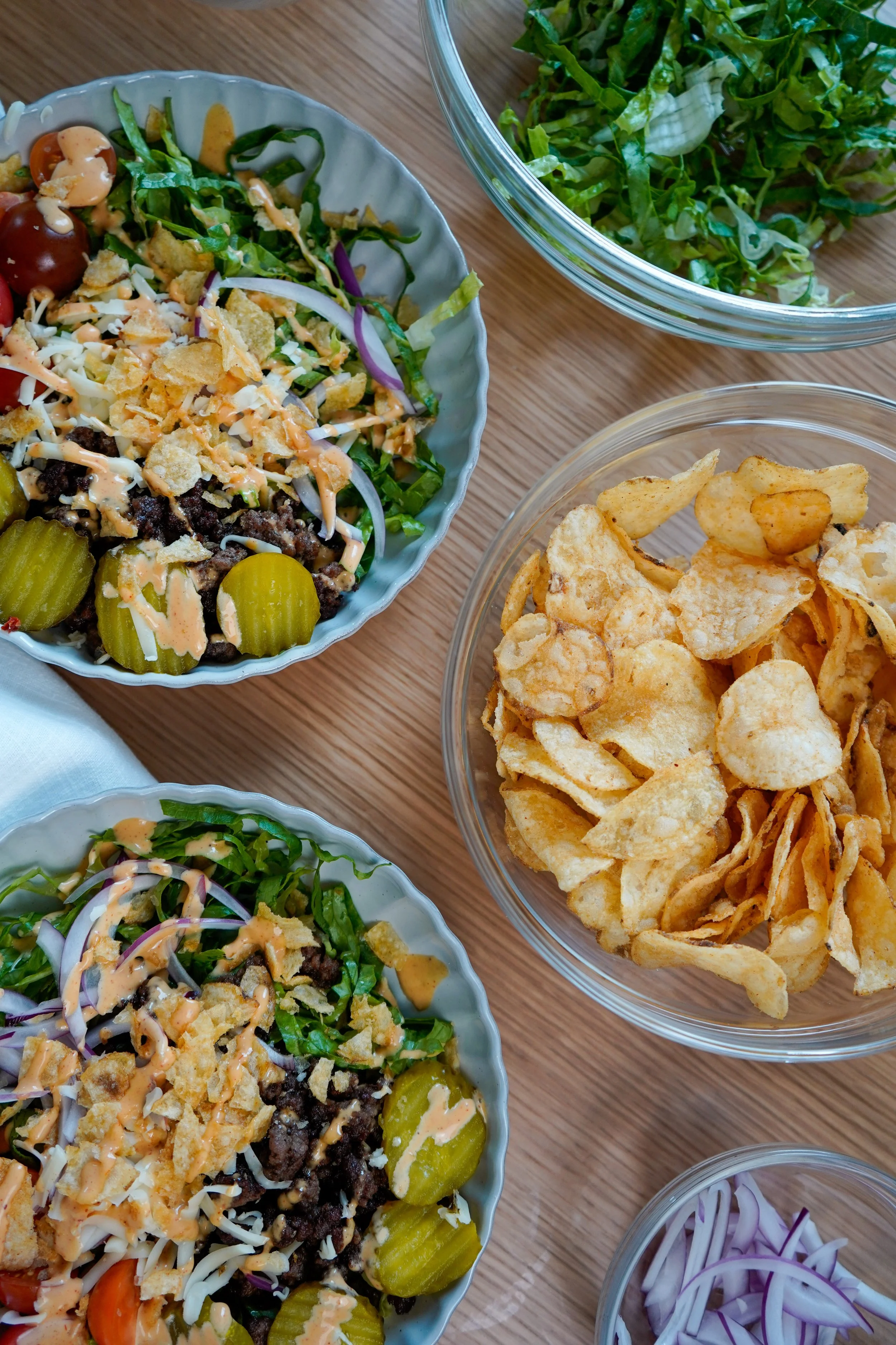 Bowls of burger bowls with shredded lettuce, tomatoes, cheese, ground beef, pickles, and tortilla chips, along with a bowl of potato chips on a wooden table.