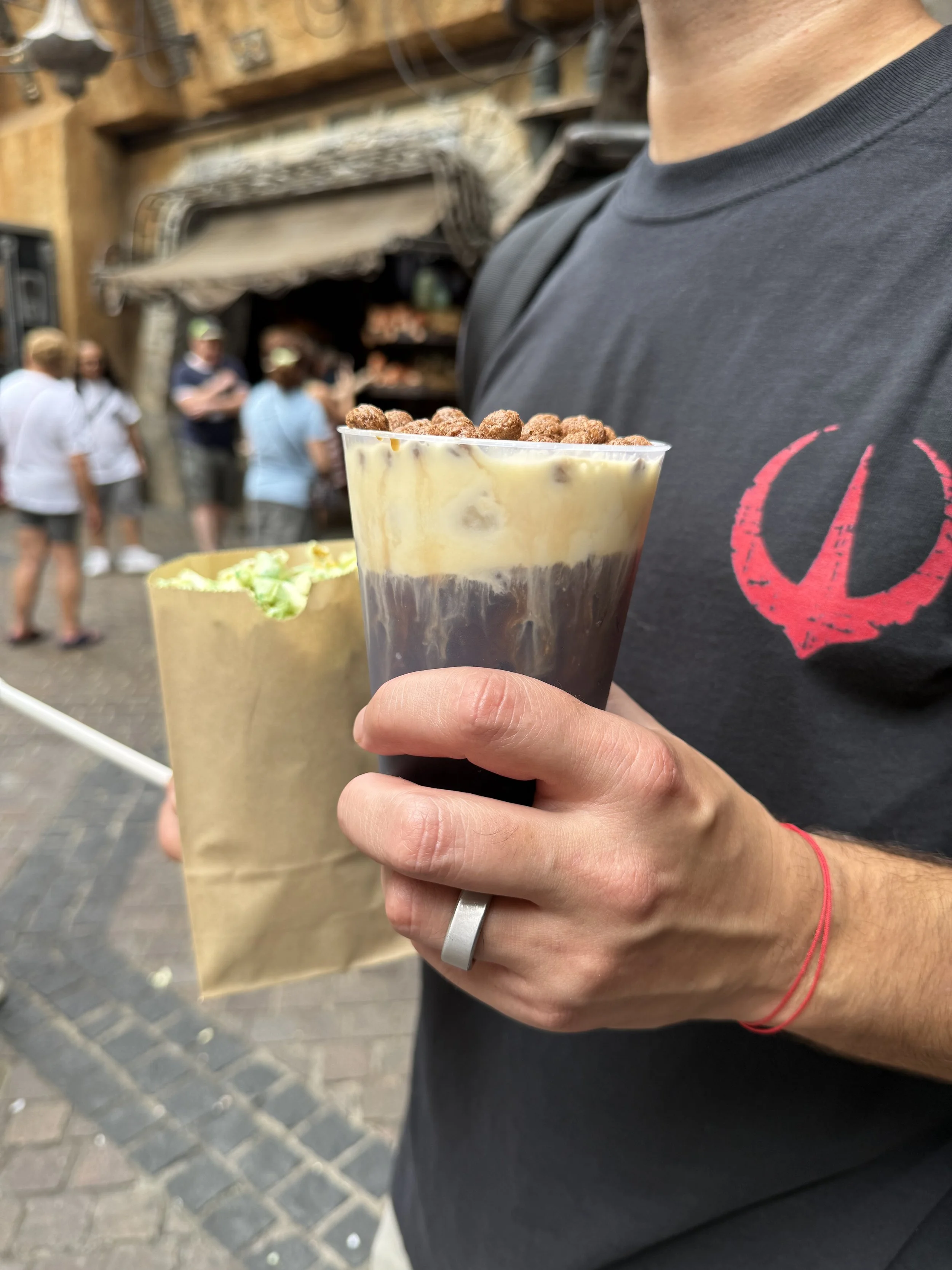 Person holding an iced coffee topped with chocolate cereal, with a paper bag containing green popcorn in the background, at Disney's Galaxy's Edge.