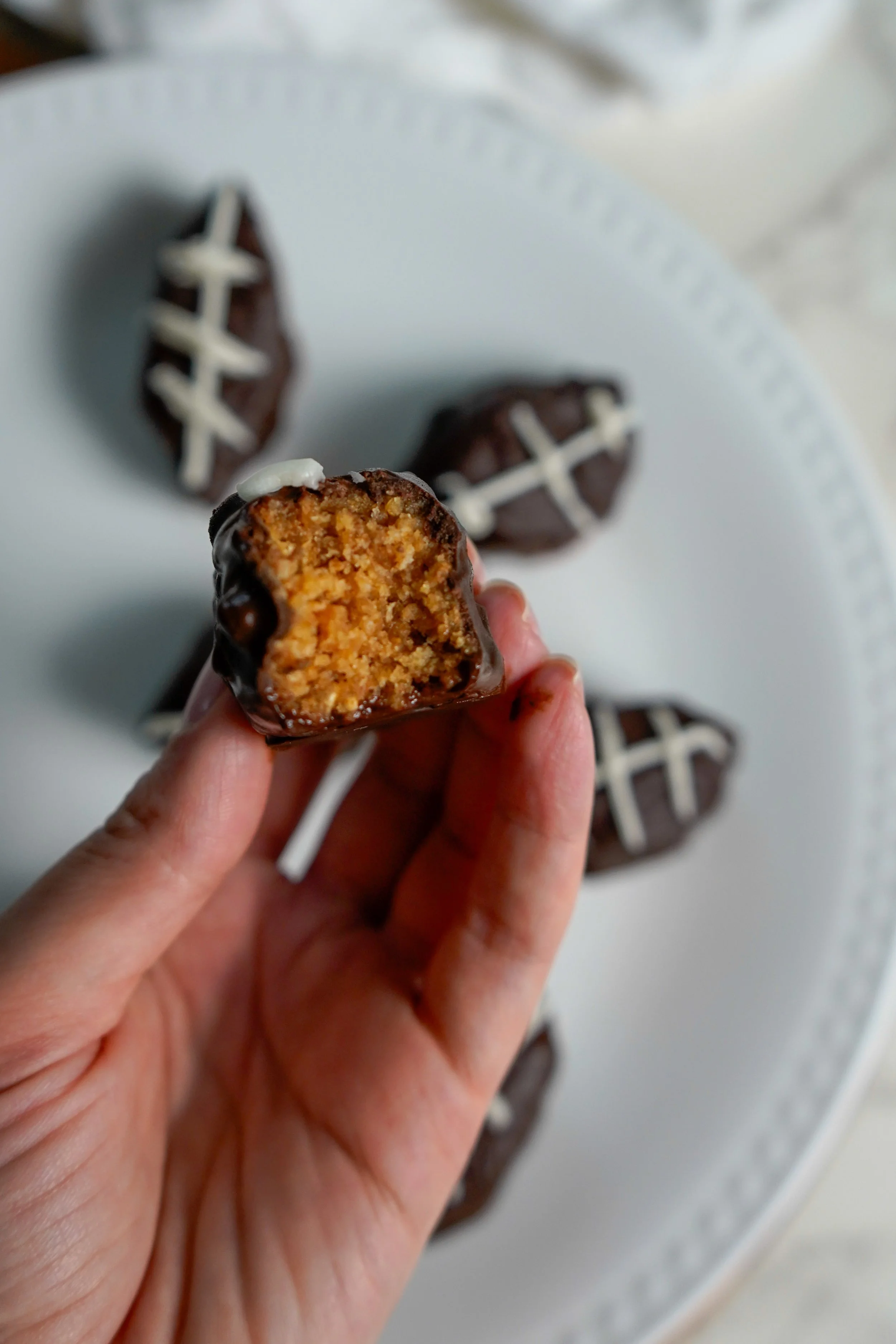 A hand holding a bitten, mini chocolate-covered snack with a visible crispy buttery pancake filling. Several similar snacks are on a white plate in the background.
