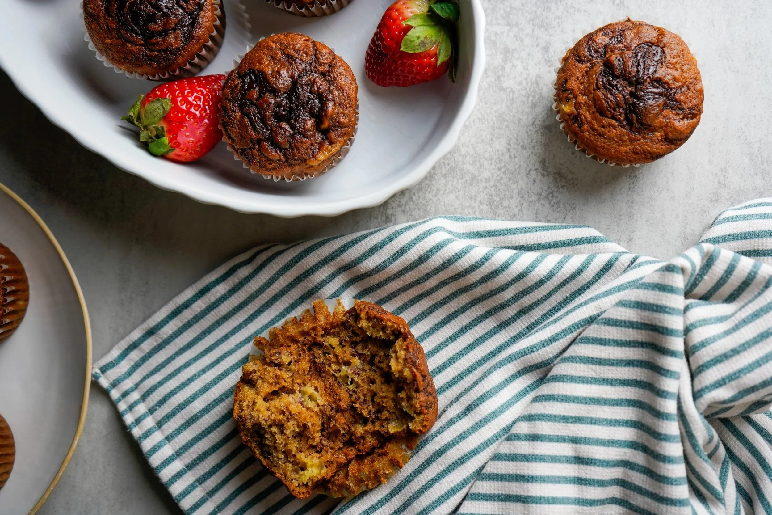Banana chocolate chip muffins on a white plate with strawberries, one muffin sliced in half revealing its interior, on a striped cloth.