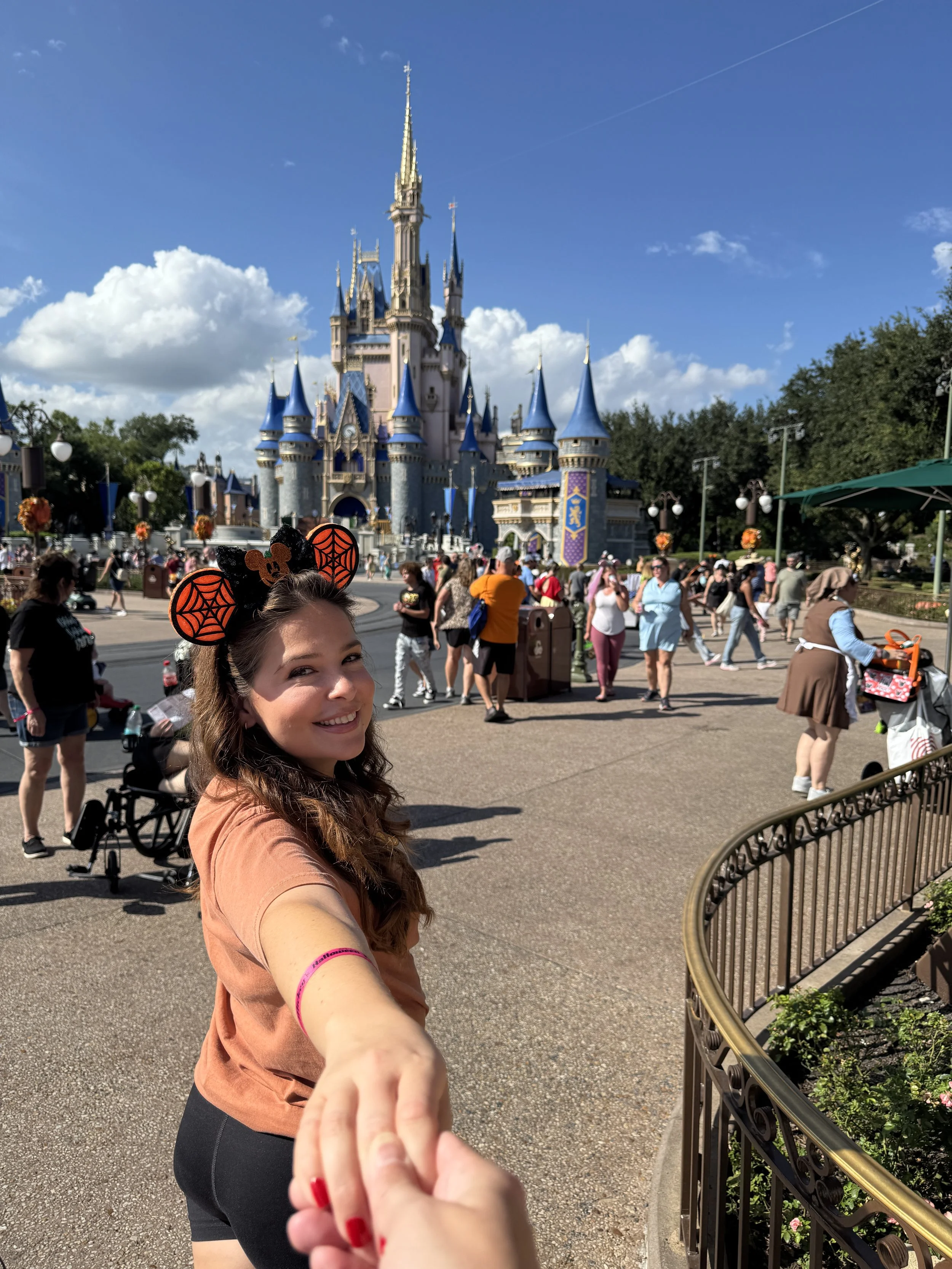 A woman in a yam colored shirt and black shorts holding hands with her husband, smiling at Disneyland with the castle in the background. She is wearing Halloween-themed ears, and the scene is busy with visitors on a sunny day.