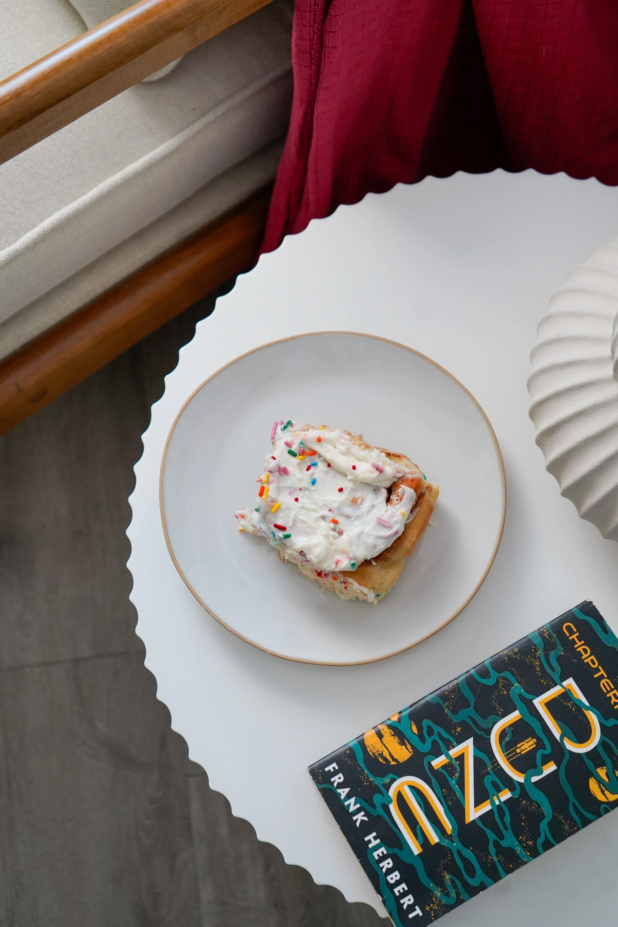 A white table with a confetti cinnamon roll with white frosting and colorful sprinkles on a white plate, a book titled 'DUNE' by Frank Herbert, and a beige textured vase, with part of a beige arm chair and pink blanket visible nearby.